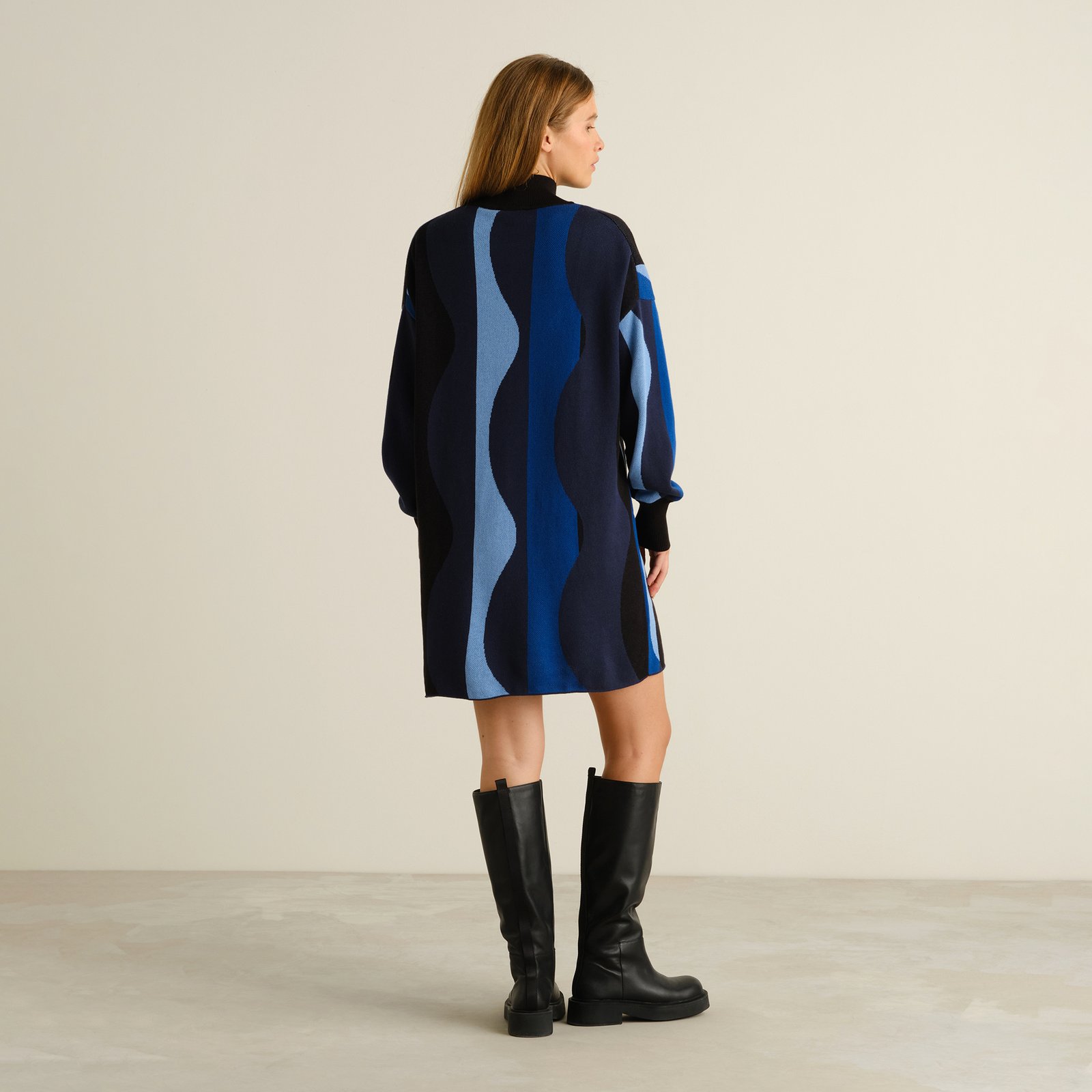 Back view of a model wearing the Nikkala dress in a blue wavy stripe pattern, styled with black knee-high boots.