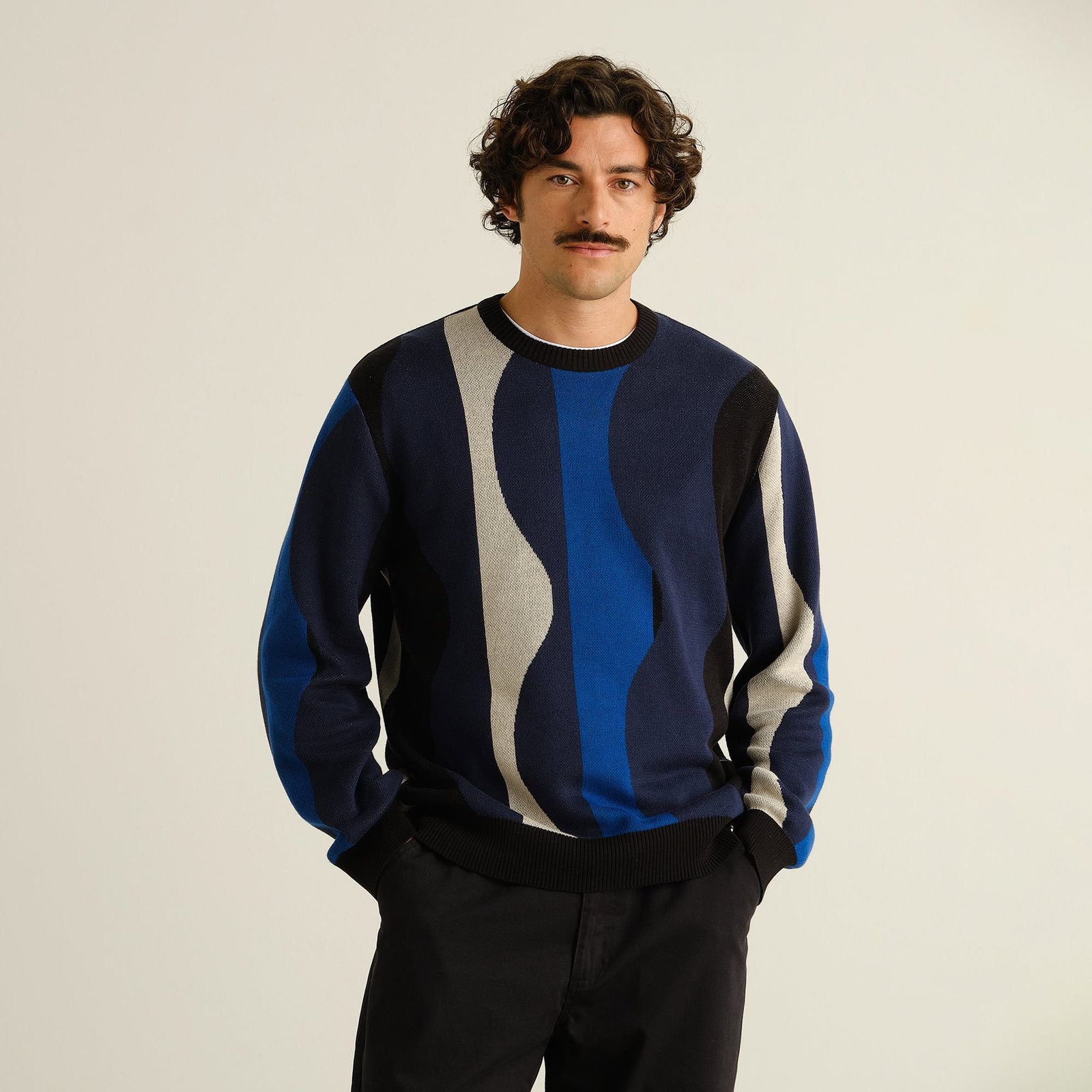 Model with a mustache wearing the Sweater Mora Wavy Stripe, posing with his hands in his pockets against a clean backdrop.