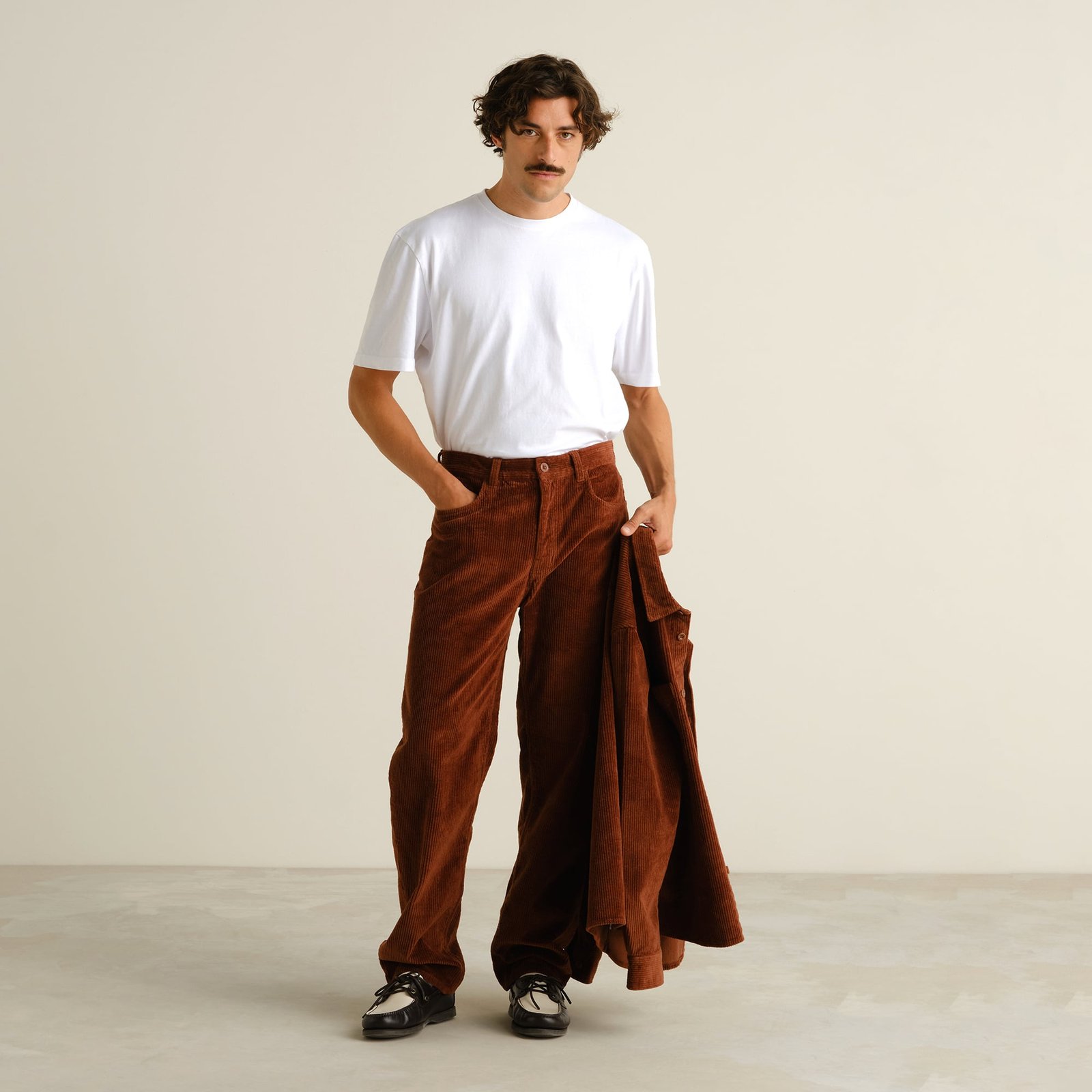 A model wears the relaxed-fit Hamburg Corduroy Pants in Cappuccino Brown with a white t-shirt, holding a matching jacket.