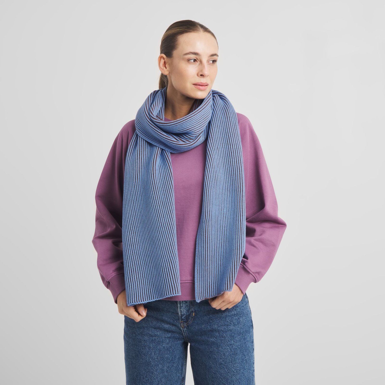 Modell som bär den grovstickade halsduken Norrfors Brioche i blått och brunt, stylad med en lila tröja och blå jeans.