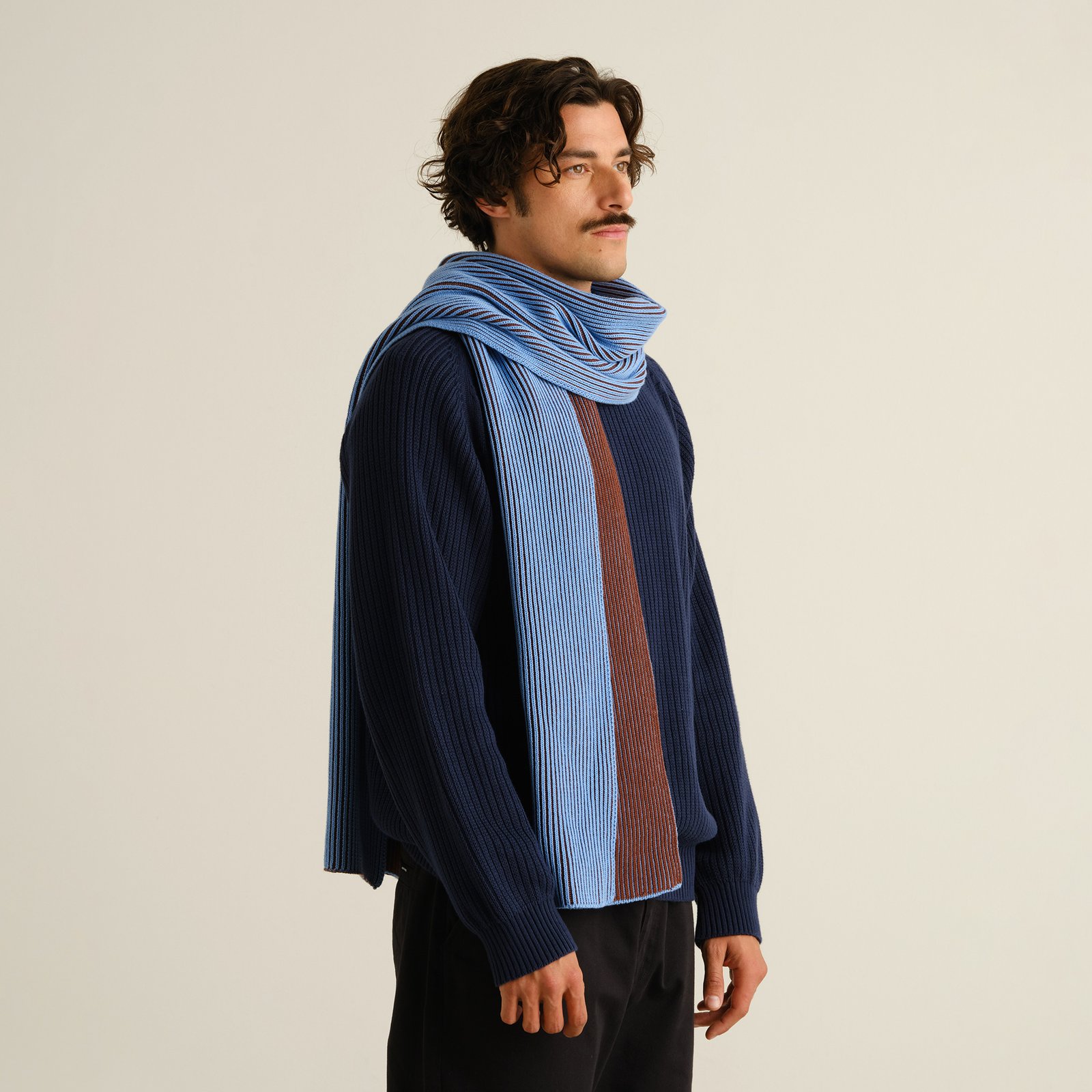 Man i profil bär den randiga halsduken Norrfors i Cappuccino Brown och Cornflower Blue till en marinblå stickad tröja.