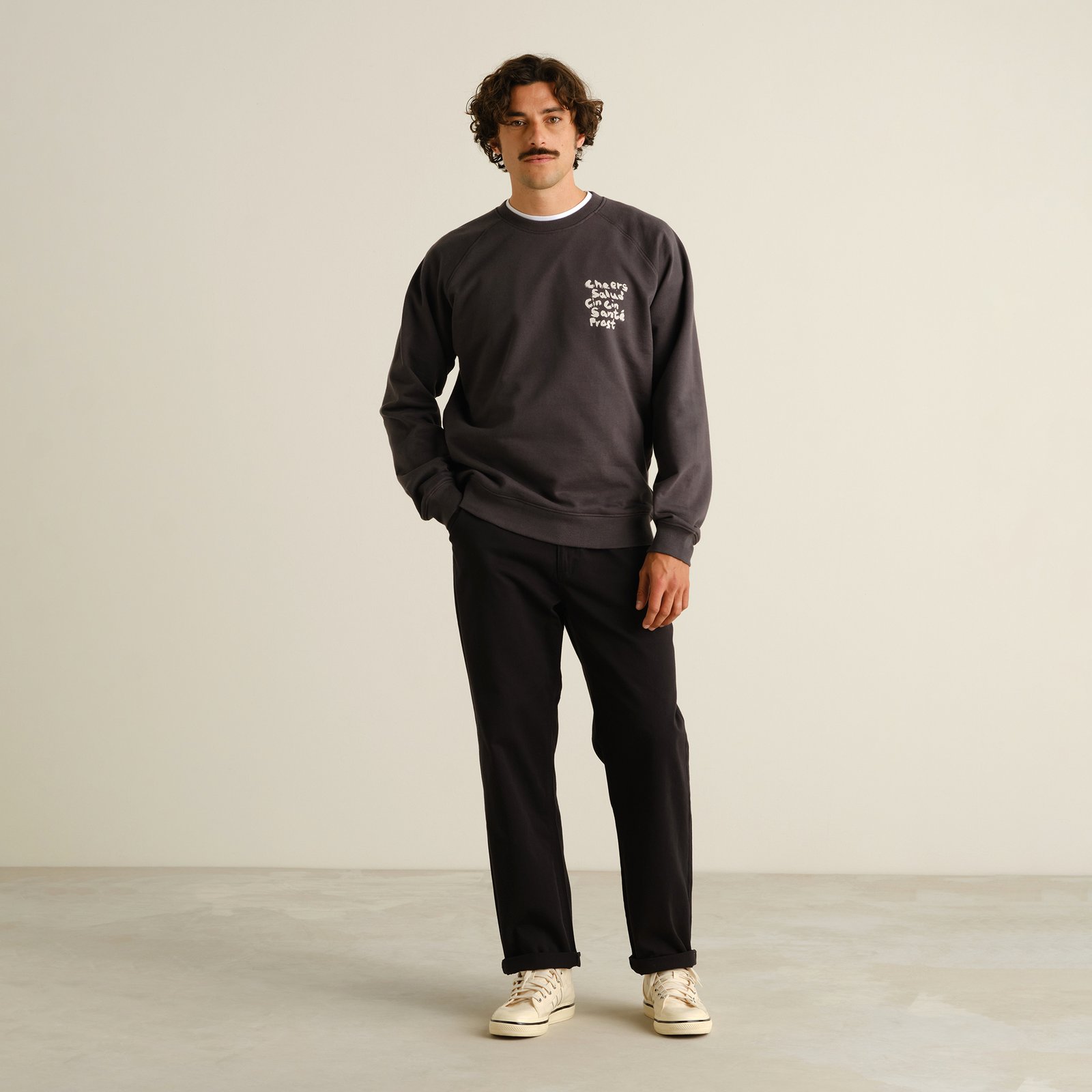 A man with a mustache wearing black Hudiksvall chinos, a dark grey sweatshirt, and off-white sneakers against a background.