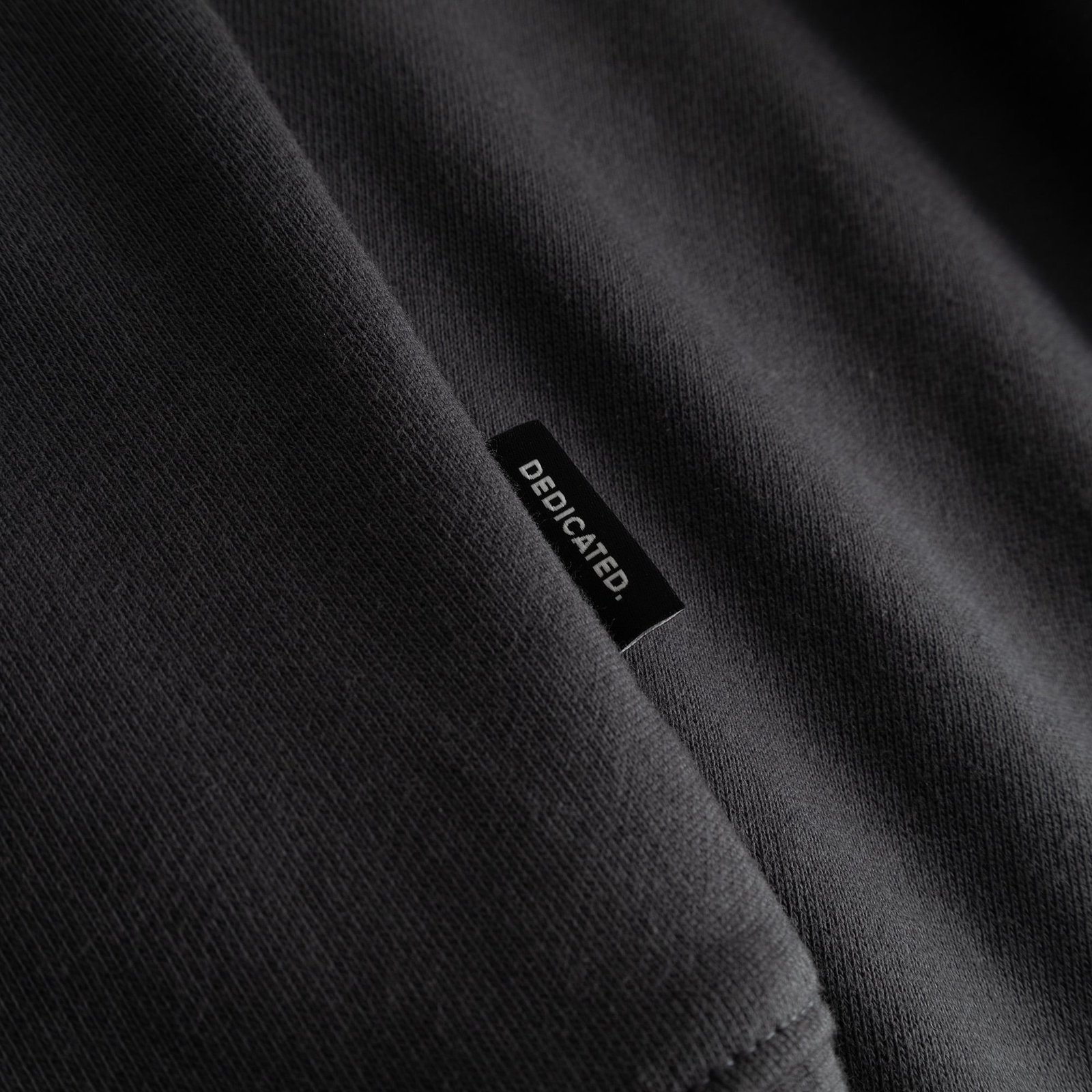 Close-up detail of the Dedicated brand label on the side of the charcoal Sweatshirt Malmoe Cheers Salud.