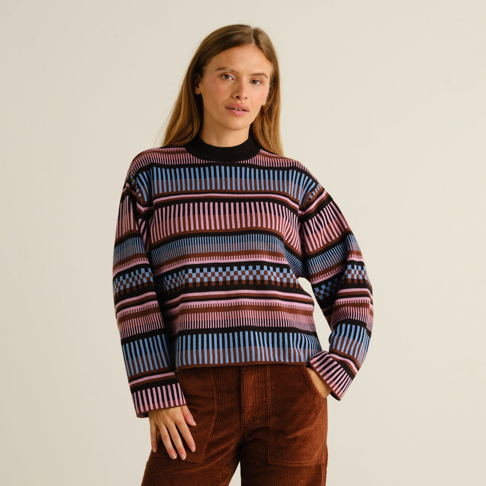 A woman models the Limhamn sweater, featuring a multi-colored dashed stripe pattern, styled with brown corduroy pants.