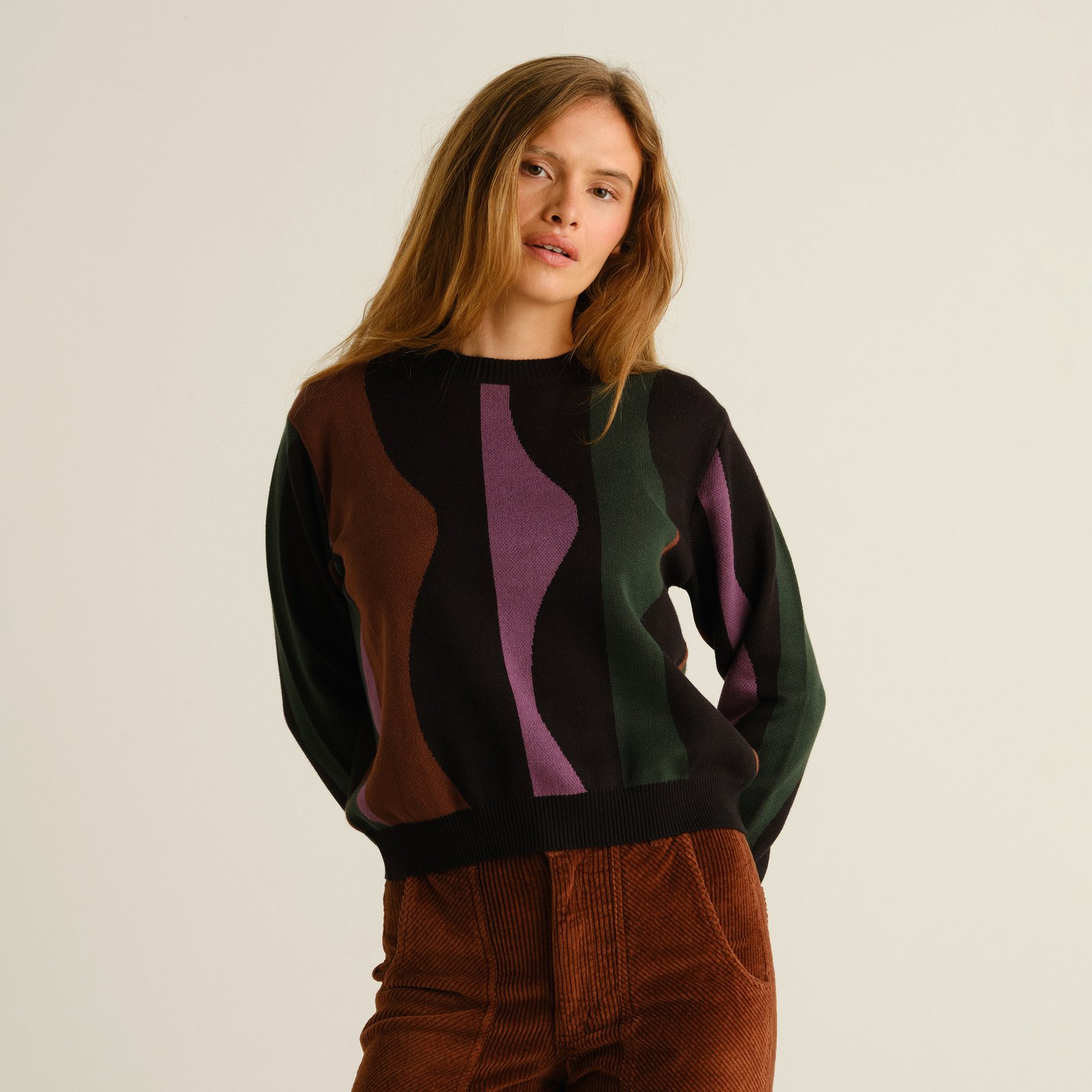 A model wears the Arendal sweater, a multi-colored knit with a wavy stripe design, paired with brown corduroy pants.