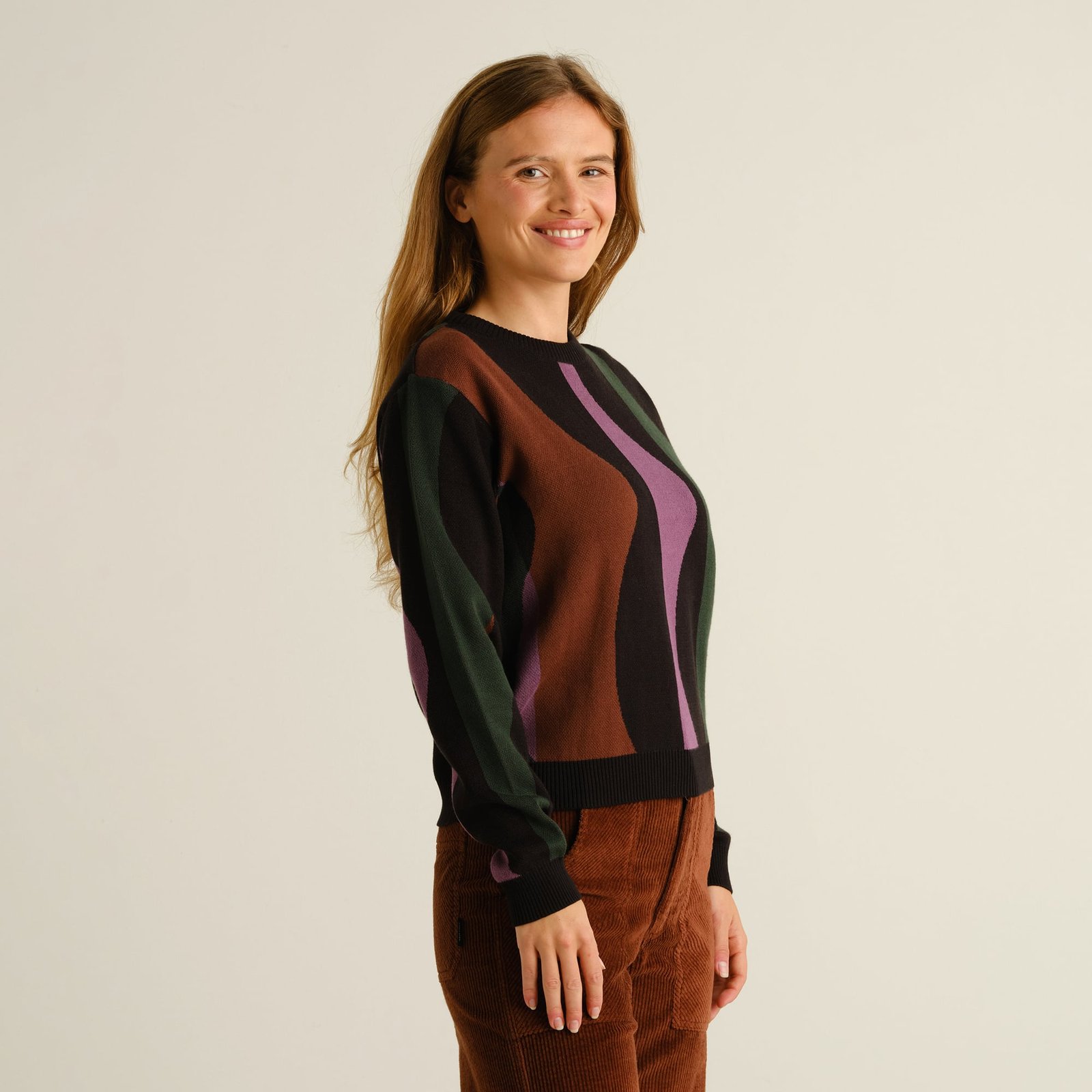 Side view of a smiling woman wearing the Sweater Arendal Wavy Stripe in Multi Color with brown corduroy trousers.