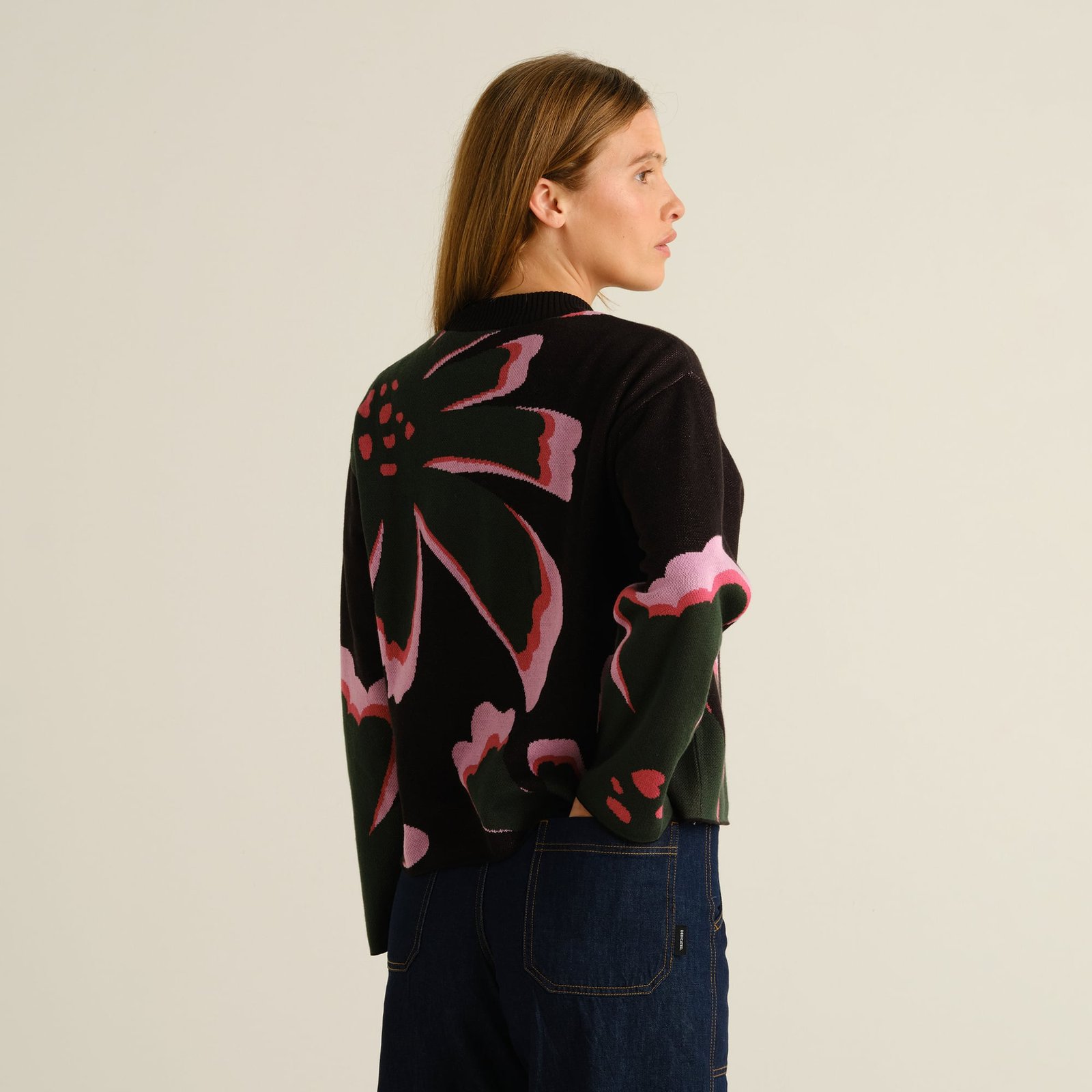A woman looks over her shoulder wearing the black Limhamn sweater with a large, colorful floral jacquard pattern on the back.