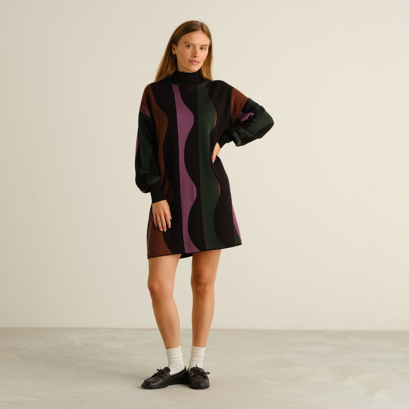 Model looking at the camera wearing Dress Nikkala, a knitted turtleneck dress with a fun multi-colored wavy stripe pattern.