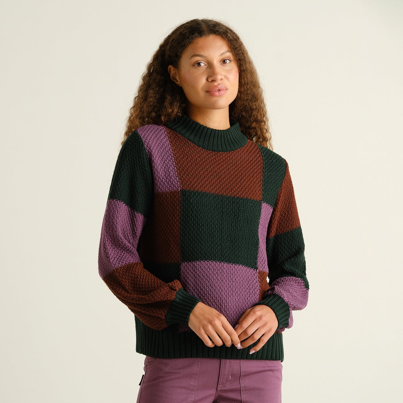 Model wears the chunky-knit Sweater Gotland, featuring a bold blocked check pattern in purple, green, and brown tones.