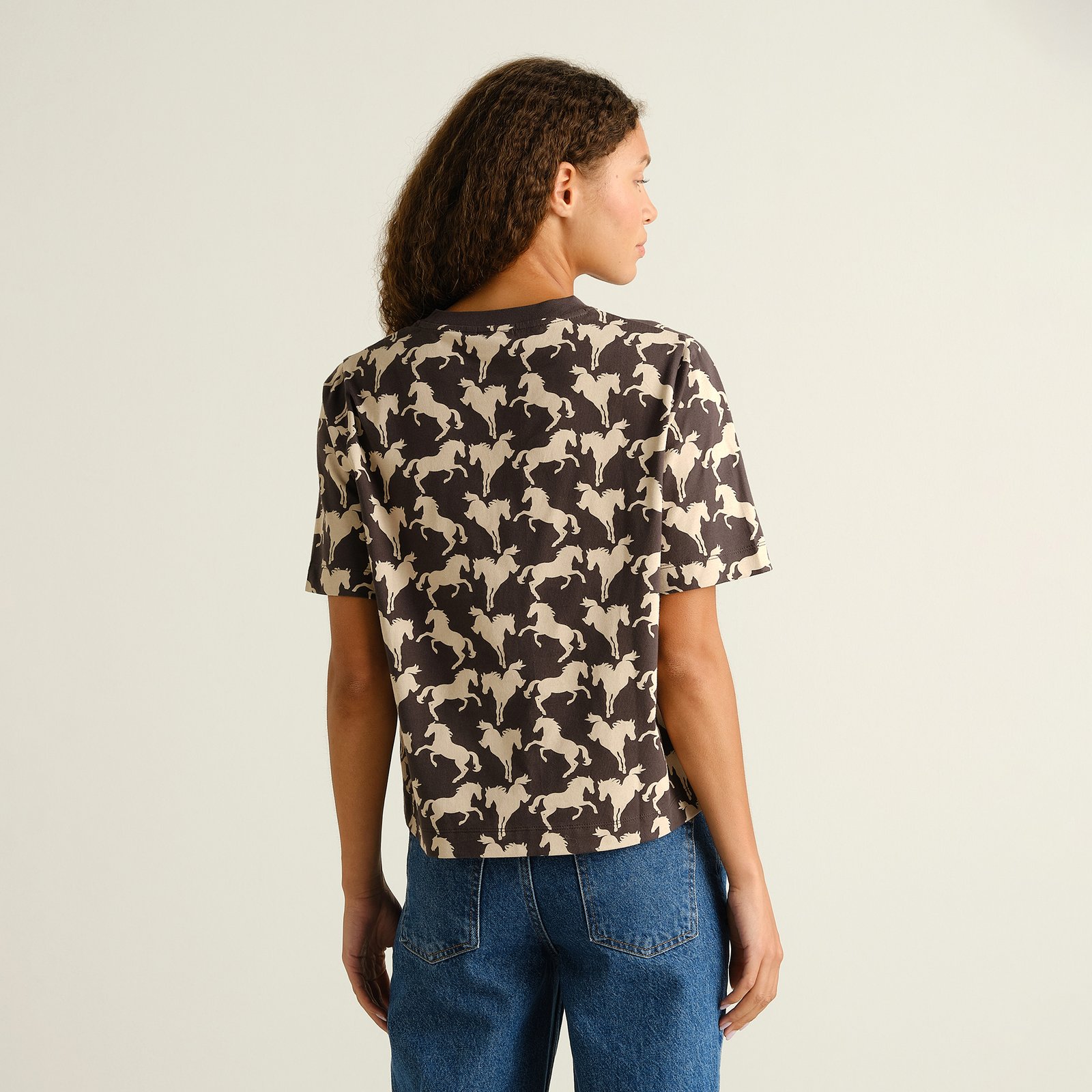 Back view of a woman wearing the charcoal T-shirt Vadstena with its all-over wild horses print, paired with blue jeans.