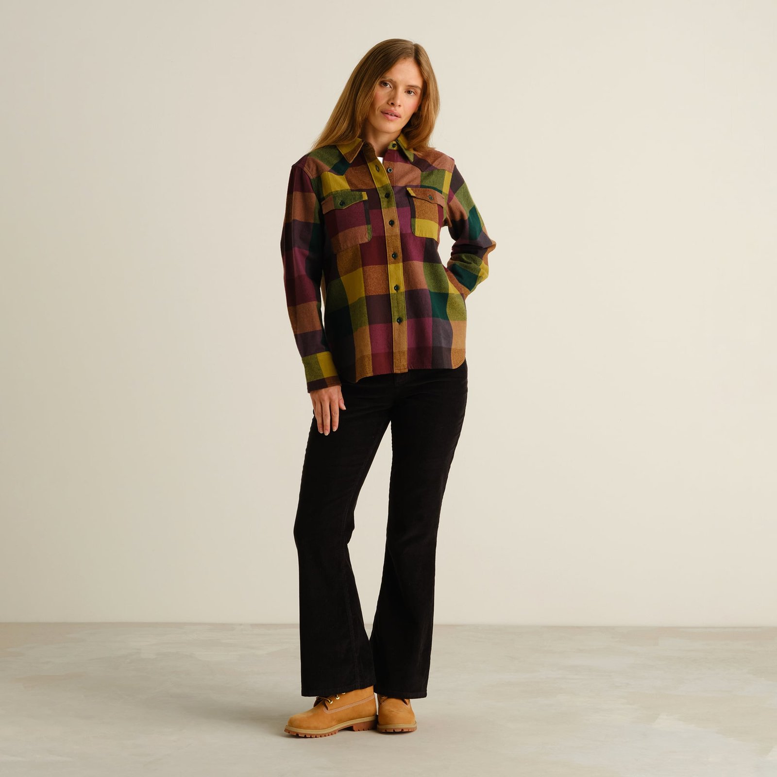 A woman models the western-inspired Arboga Flannel Shirt in a bold multicolor check with black corduroy pants and tan boots.