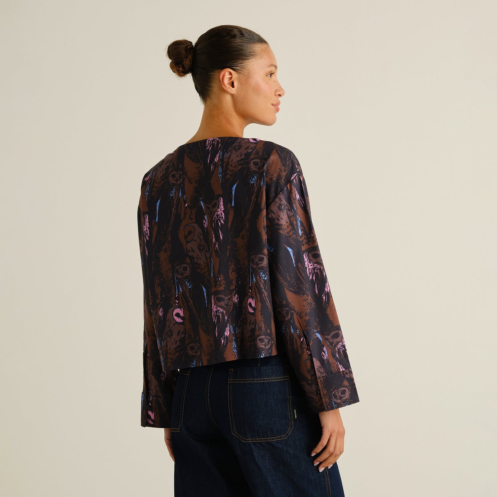 A woman with her hair in a bun shows the back of the Top Udda, a boxy blouse with a vibrant owl print.