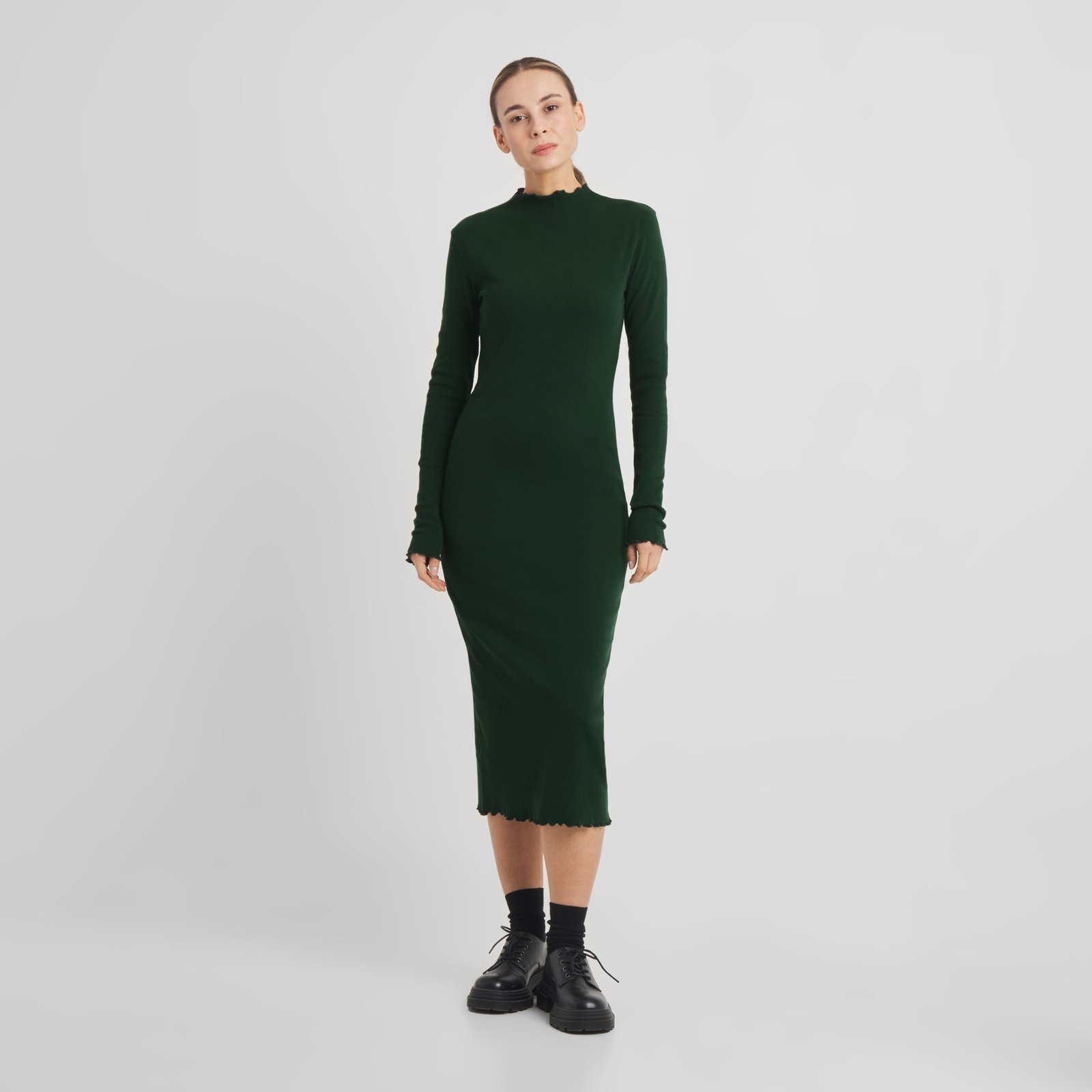 Model wearing the long-sleeved Rib Dress Lofta in dark green, styled with black socks and chunky black shoes.