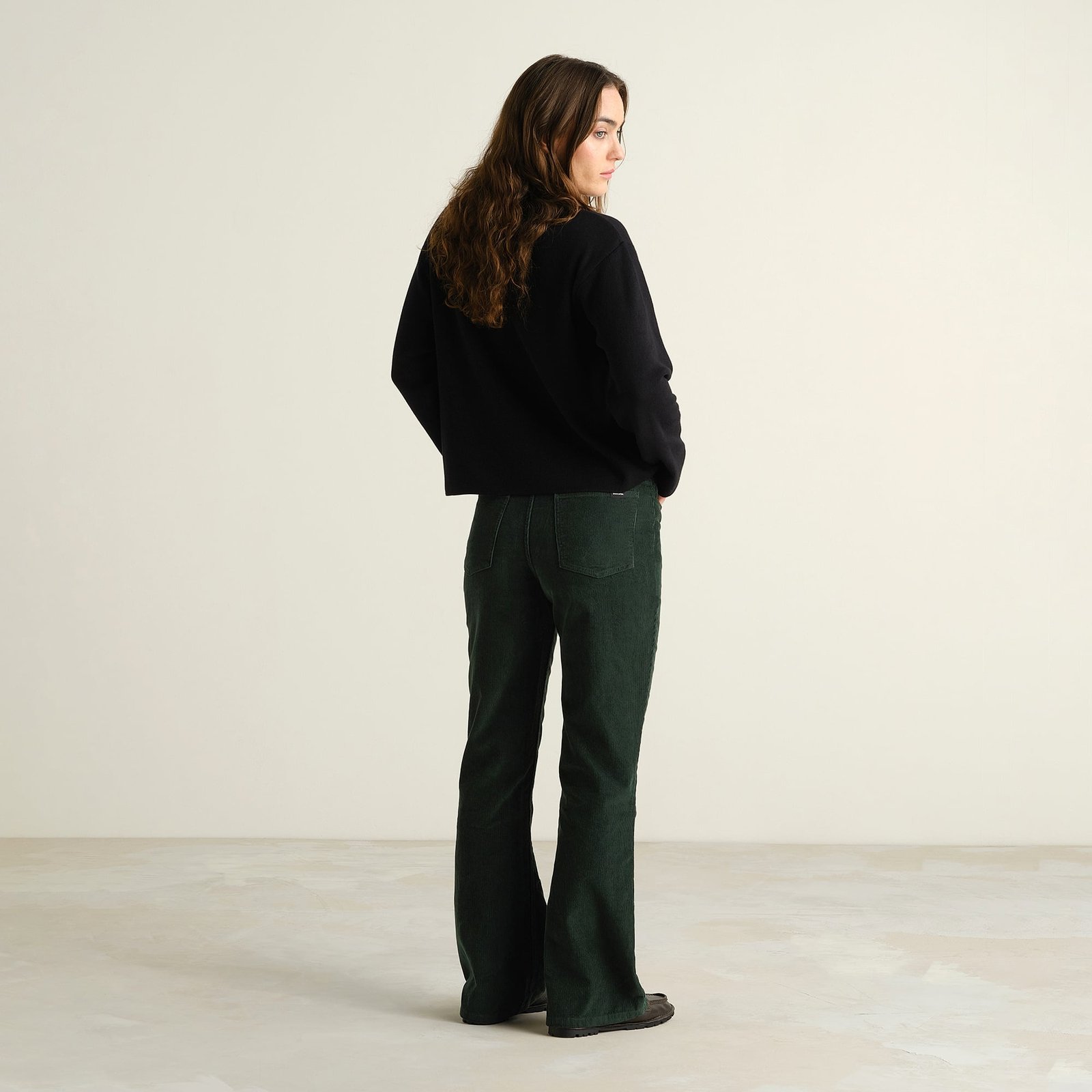 Woman looks over her shoulder while wearing the Ryda Corduroy Pants in Dark Green with a black sweater.