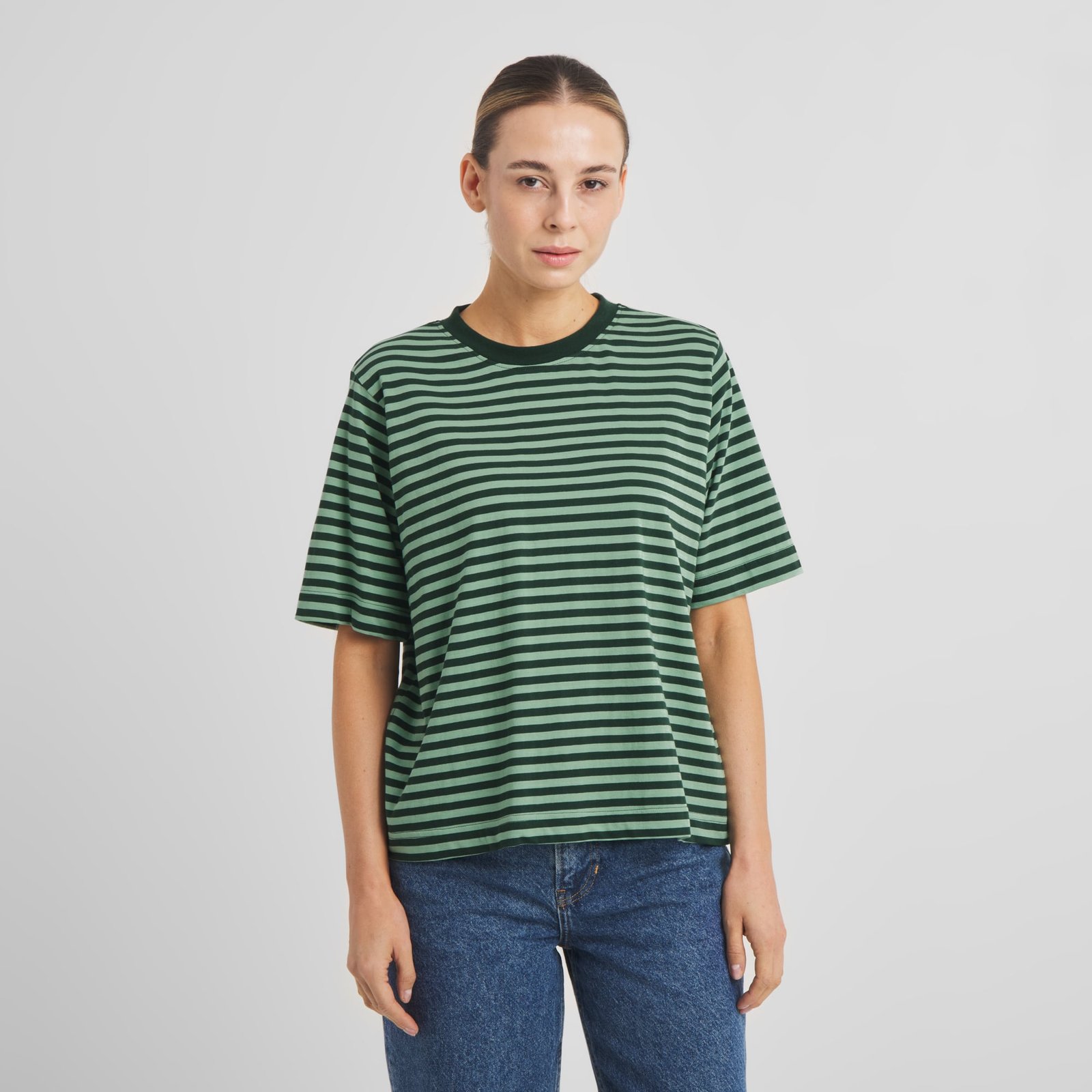 A woman with her hair pulled back models the relaxed-fit tee Vadstena in Dark Green/Granite Green stripes with blue jeans.