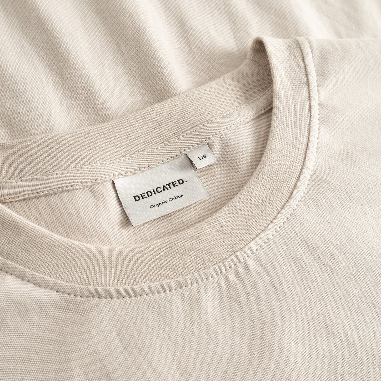 Close-up of the Dedicated brand label and ribbed collar on the classic Stockholm t-shirt in a soft oat white.