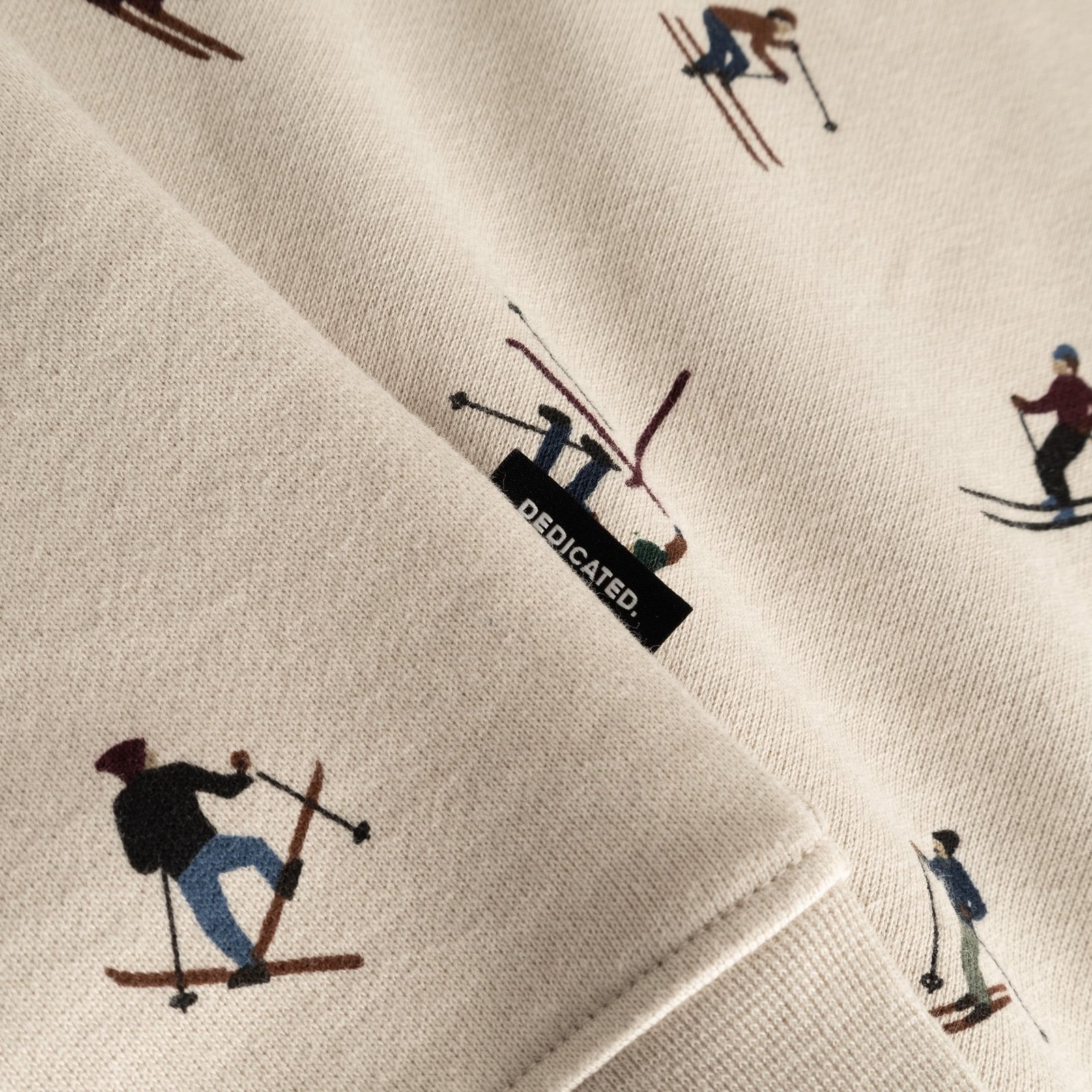 Close-up detail shot of the oat white Malmoe Ski Team sweatshirt fabric, showing the fun skier print and Dedicated brand tab.