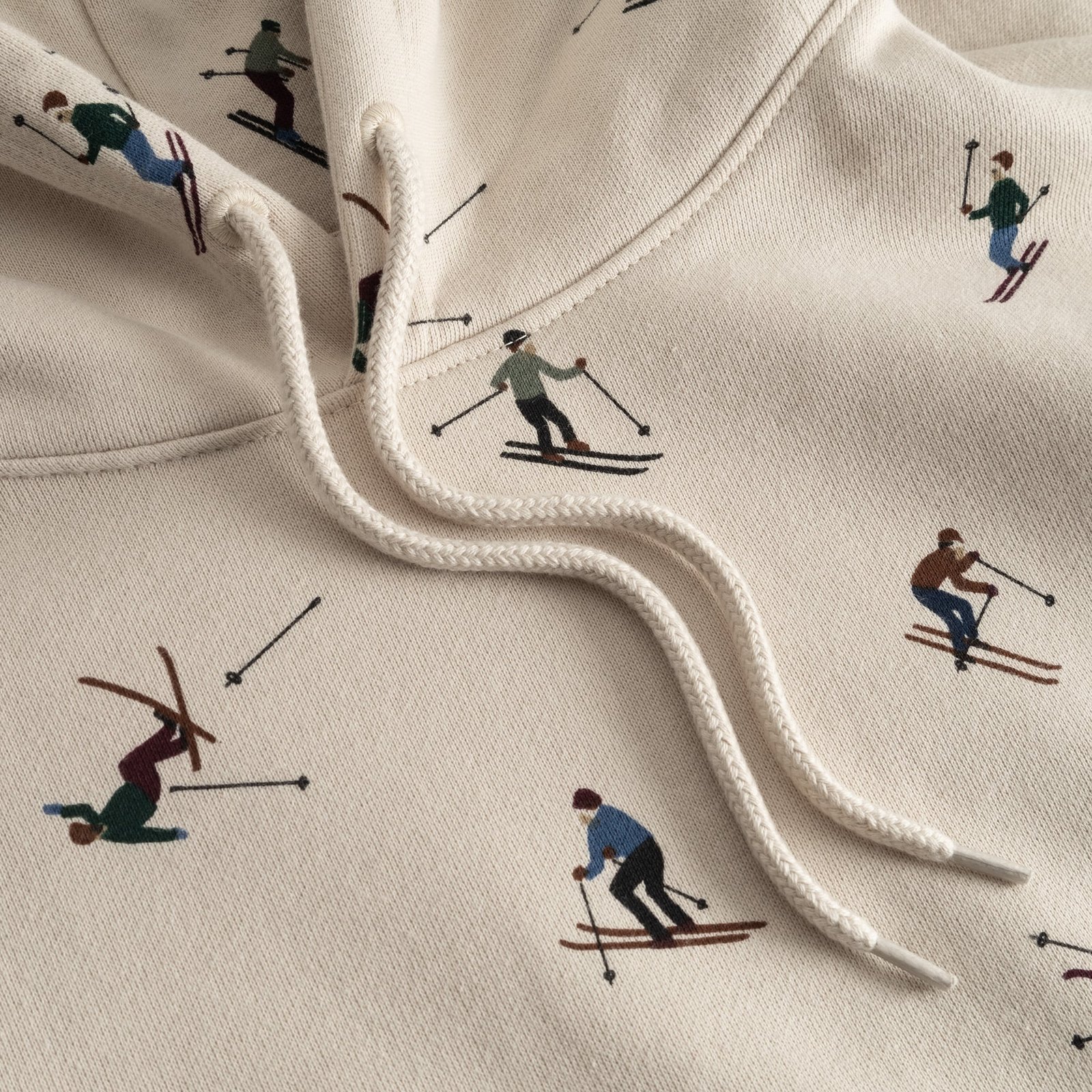 Close-up detail of the oat white Falun Ski Team hoodie, showing the playful all-over embroidered skier print and drawstrings.