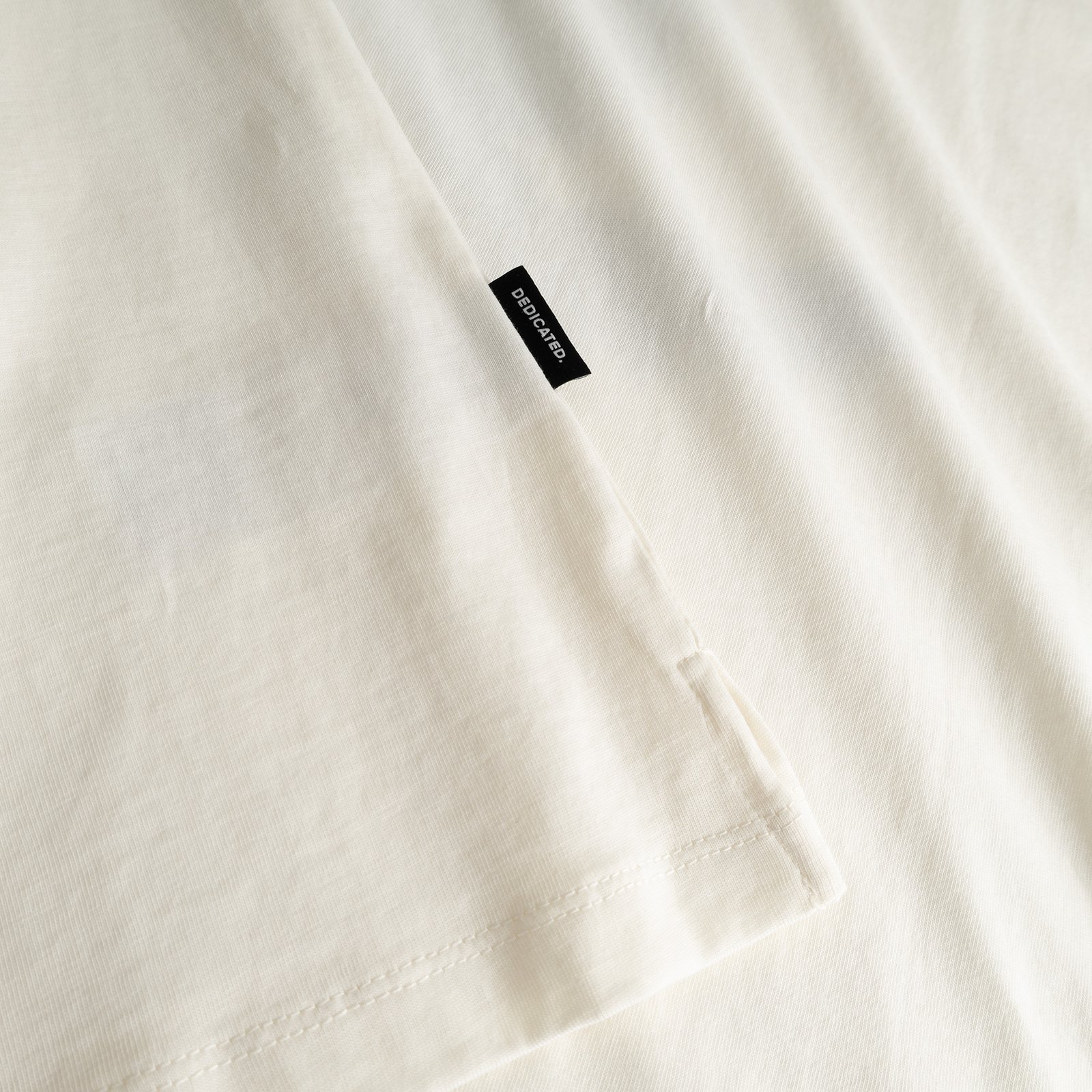 Close-up of the off-white T-shirt Visby, showing the organic cotton fabric, side slit, and a small Dedicated brand tab.