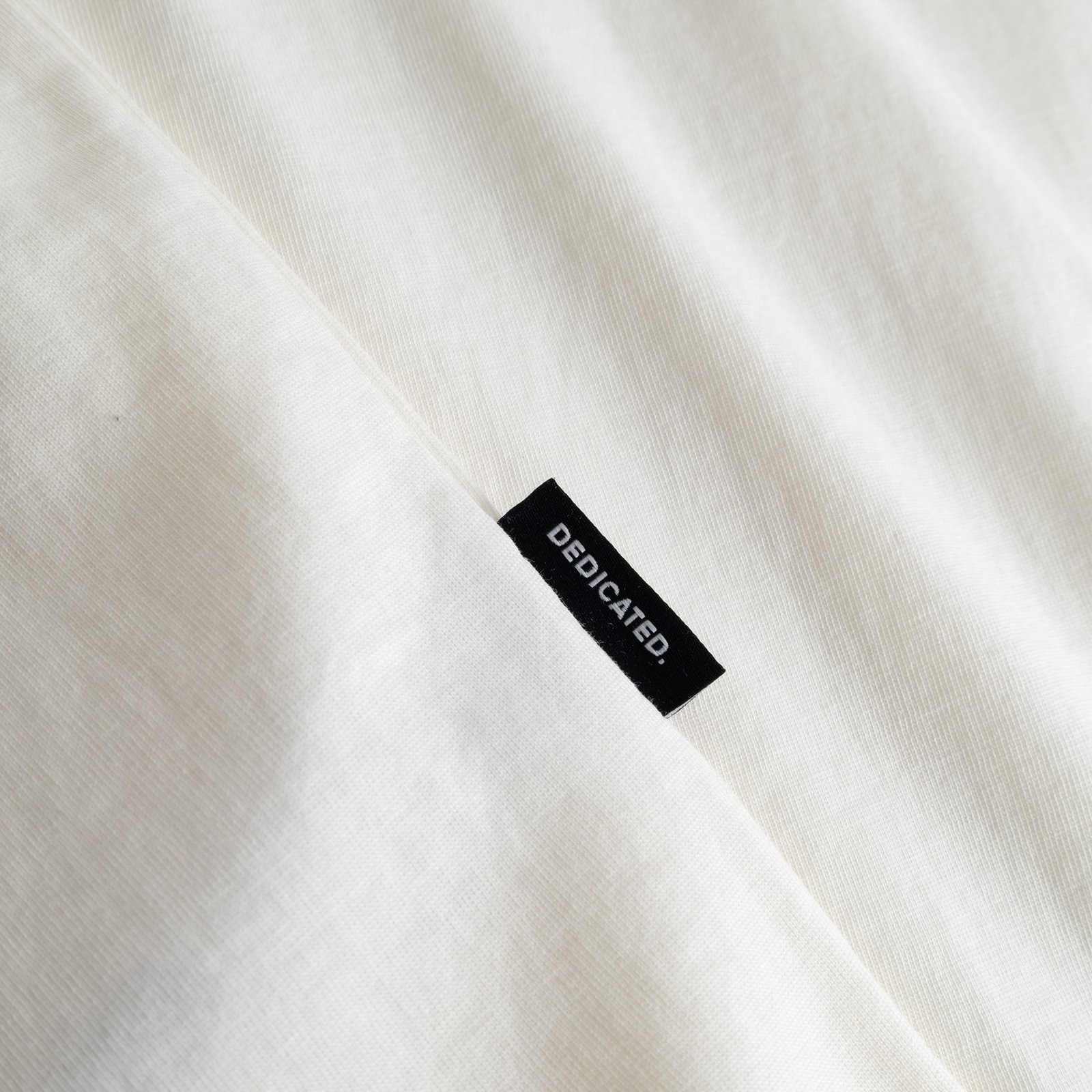 Close-up of the black Dedicated brand tab on the side seam of the off-white T-shirt Stockholm Camp Crew.