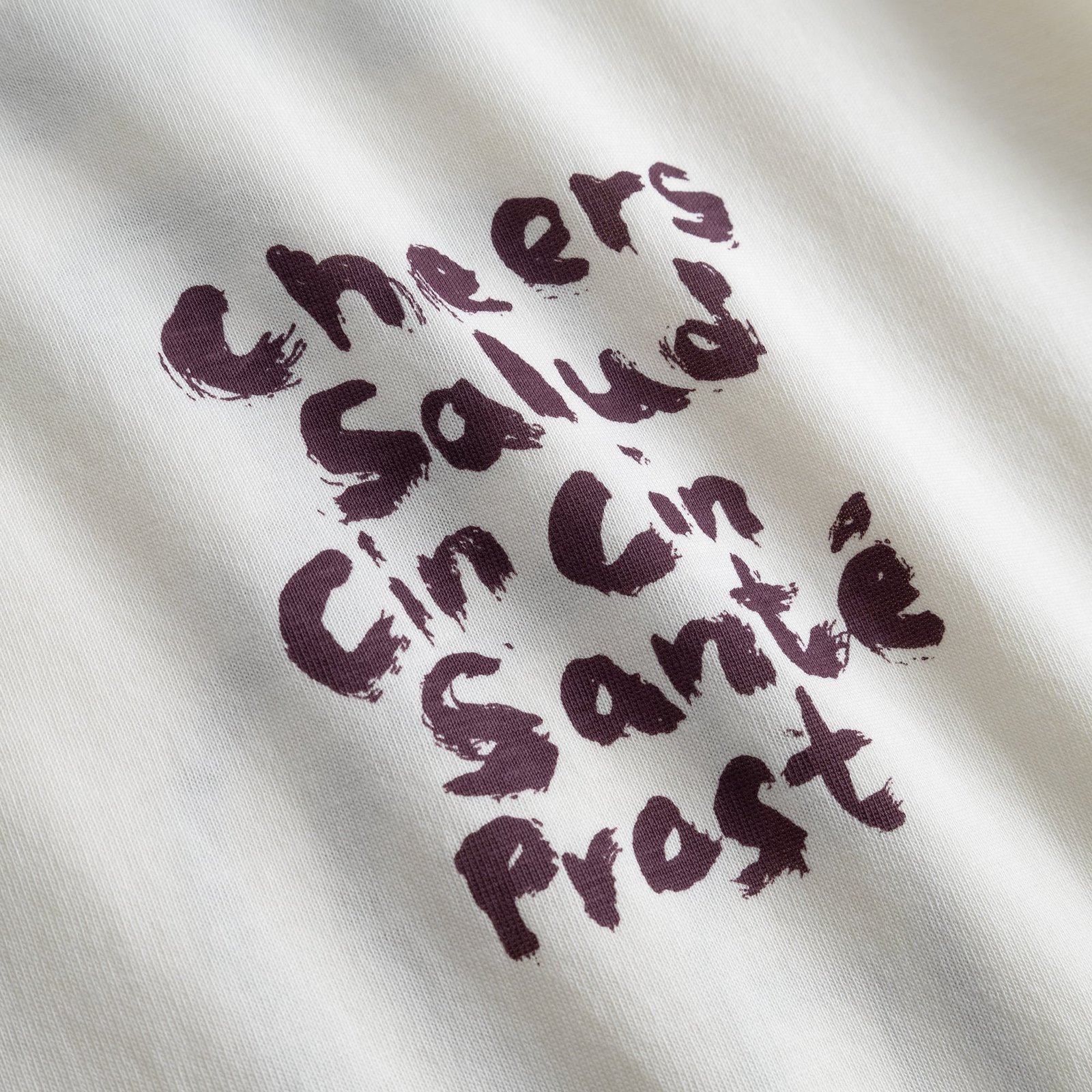Nahaufnahme des dunkelvioletten Brustaufdrucks „Cheers Salud“ im Pinselstrich-Stil auf dem T-Shirt Stockholm.