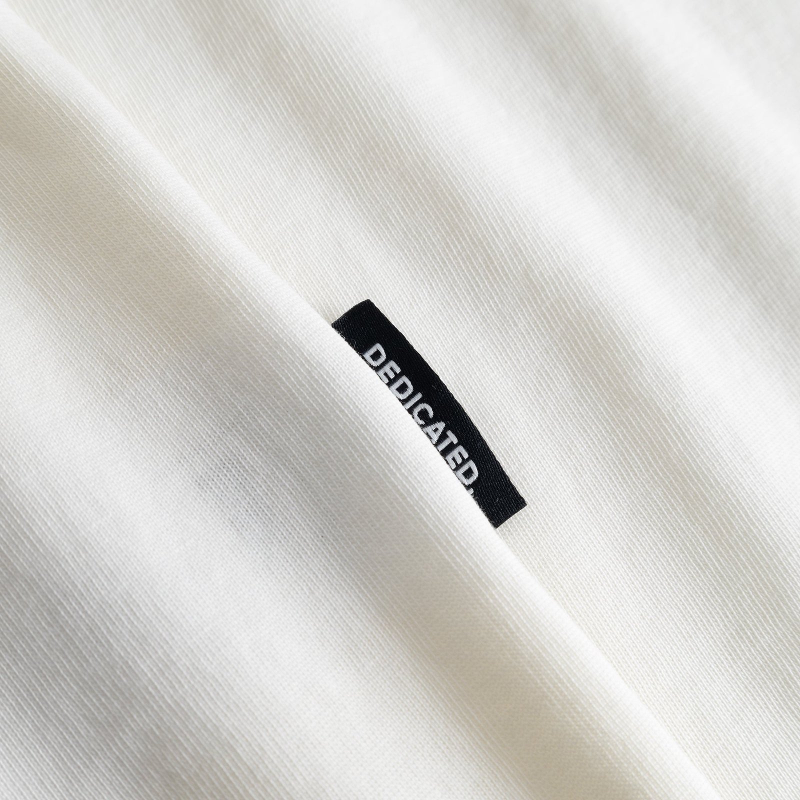 Nahaufnahme des klassischen, schwarzen Dedicated-Labels auf dem weichen, cremefarbenen T-Shirt Stockholm Cheers Salud.