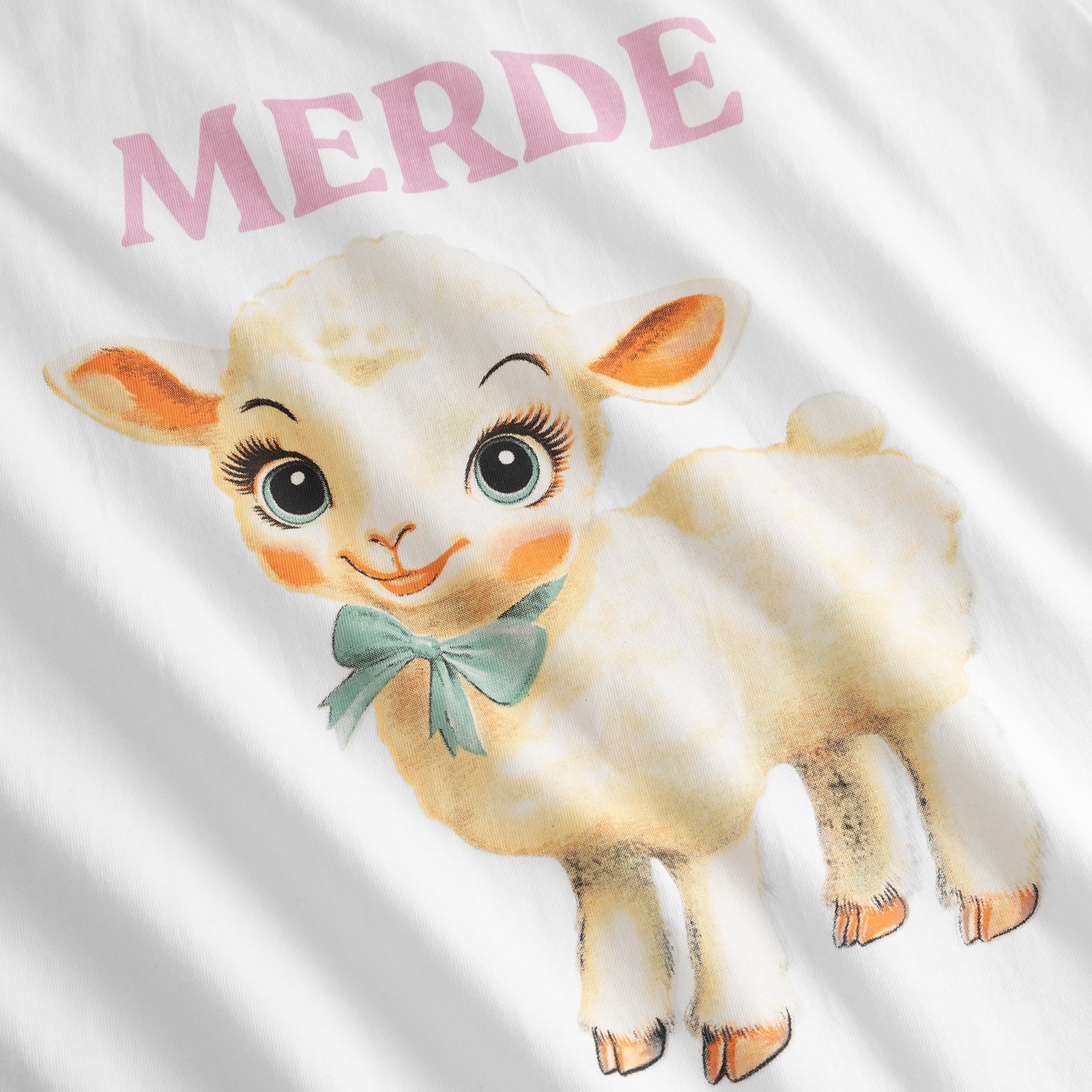 A close-up of the T-shirt Vadstena in white, showcasing its fun vintage-style lamb graphic and the word 'Merde'.