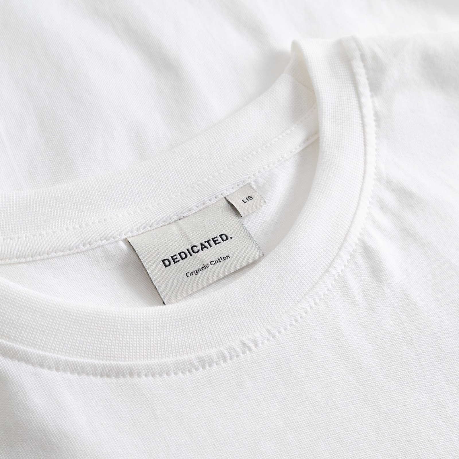 Close-up of the Dedicated brand label and ribbed collar on the white Stockholm Camp Crew t-shirt.