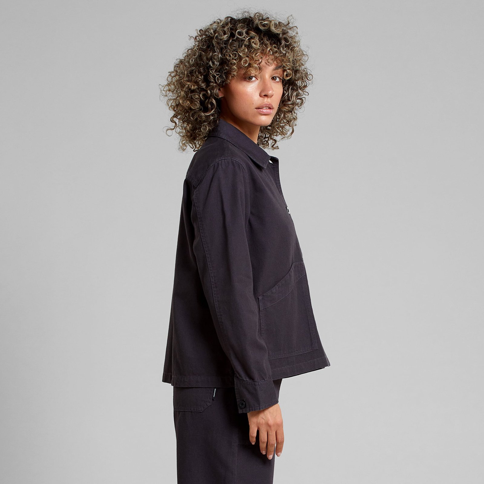 A model with curly hair looks over her shoulder, showing the side view of the Stiby Canvas Jacket in Phantom Black.