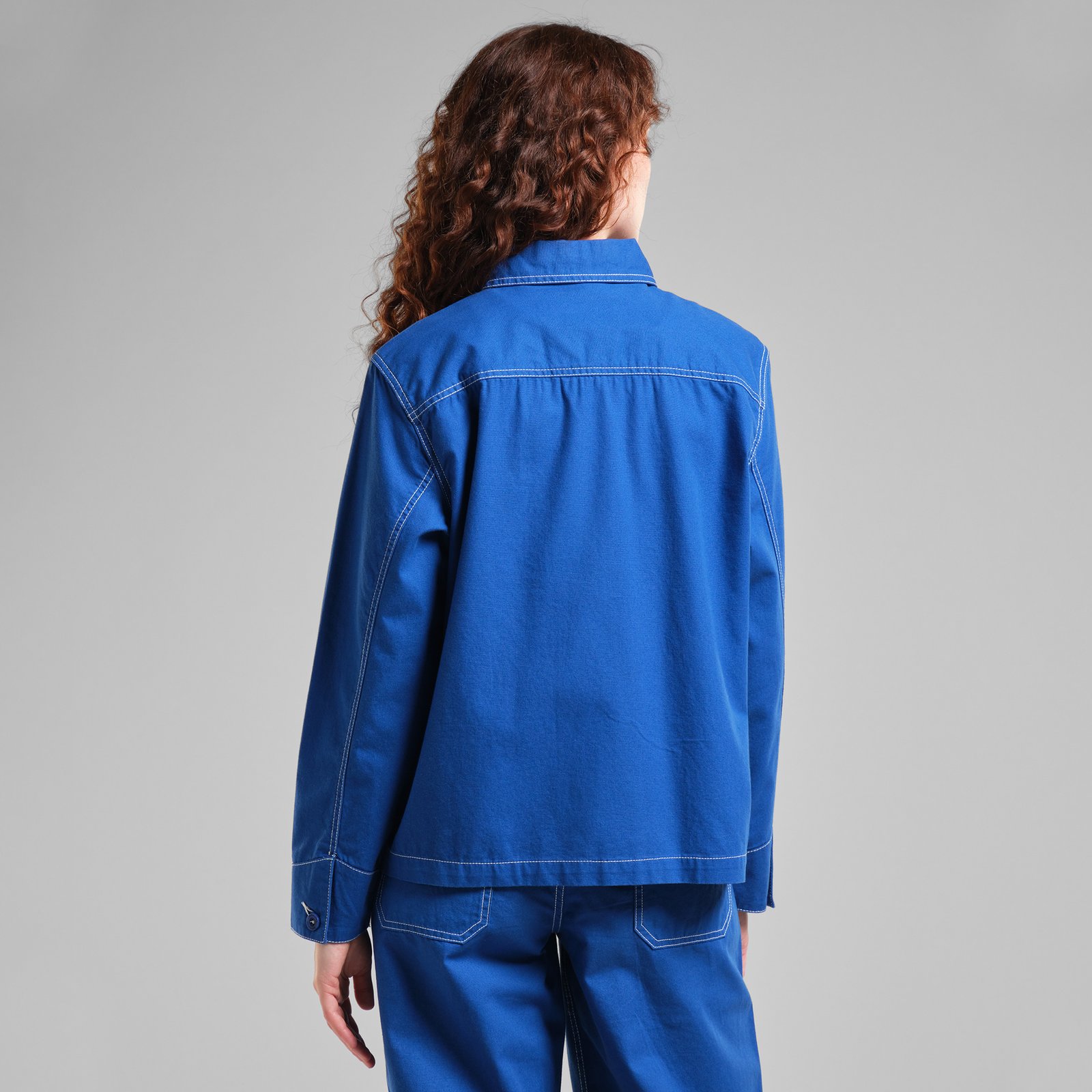 Back view of a model wearing the bright blue Stiby Canvas Jacket with matching trousers against a grey studio background.