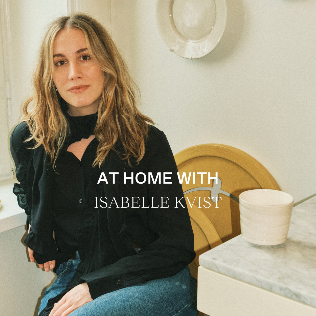 Interior featuring a round table with a linen tablecloth, set with colorful Mateus ceramic bowls and plates in red, yellow, and plum. A vase with poppies serves as a centerpiece, with the text 'AT HOME WITH' overlaying the scene.