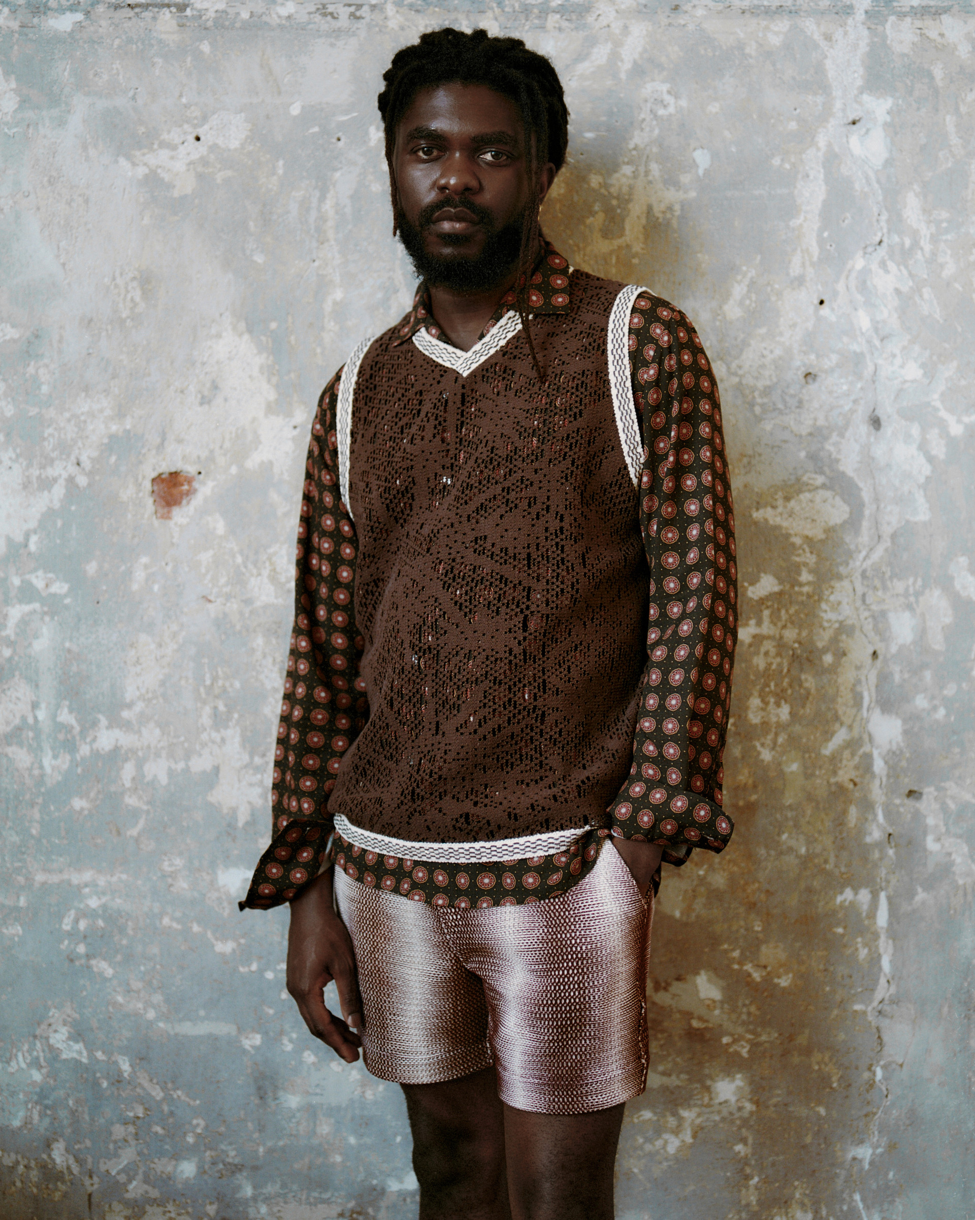 Model in a textured brown vest with trim details over a printed shirt, and gradient swim shorts against a textured wall.