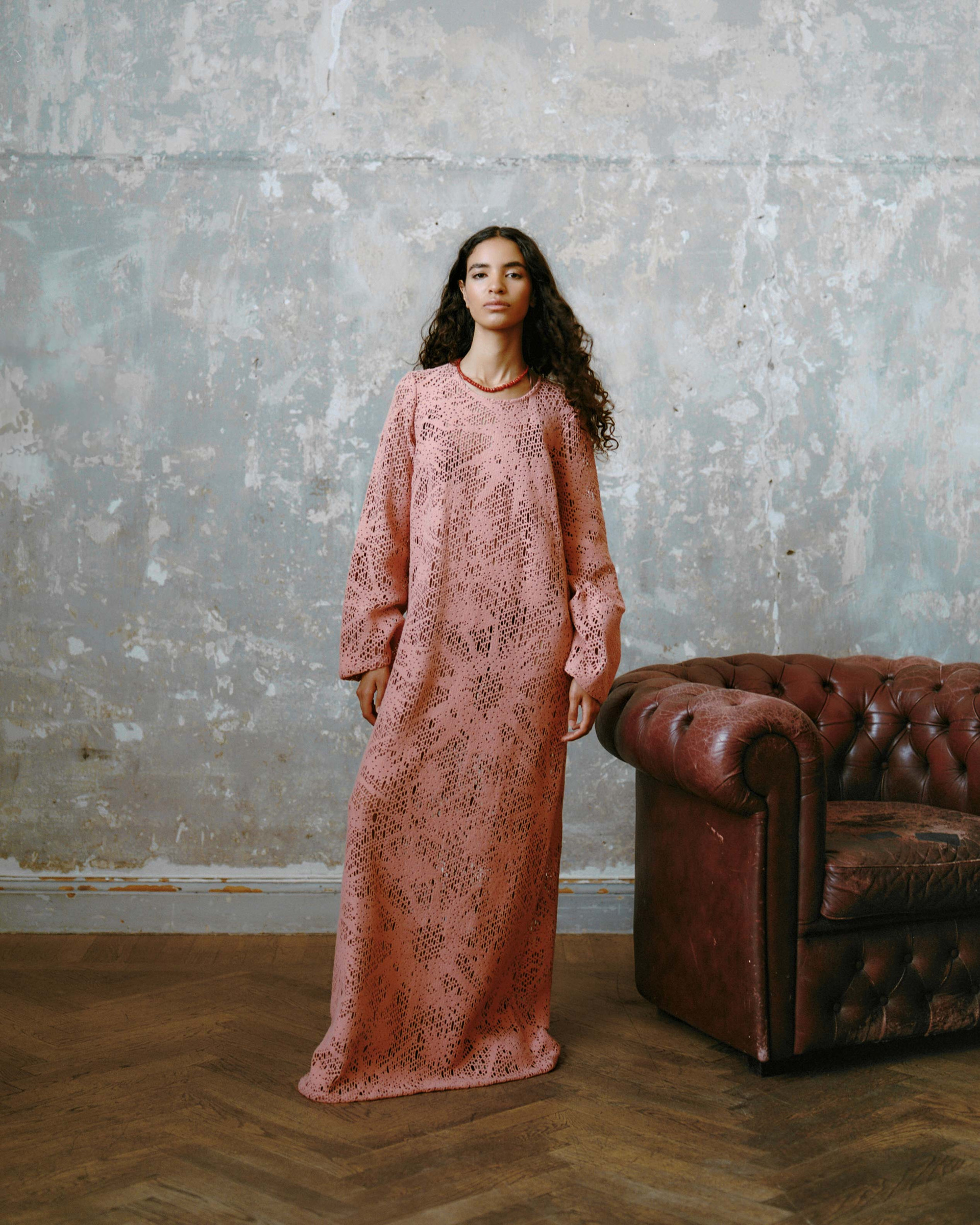Model in a full-length crochet dress in dusty pink, paired with a beaded necklace against a textured wall.