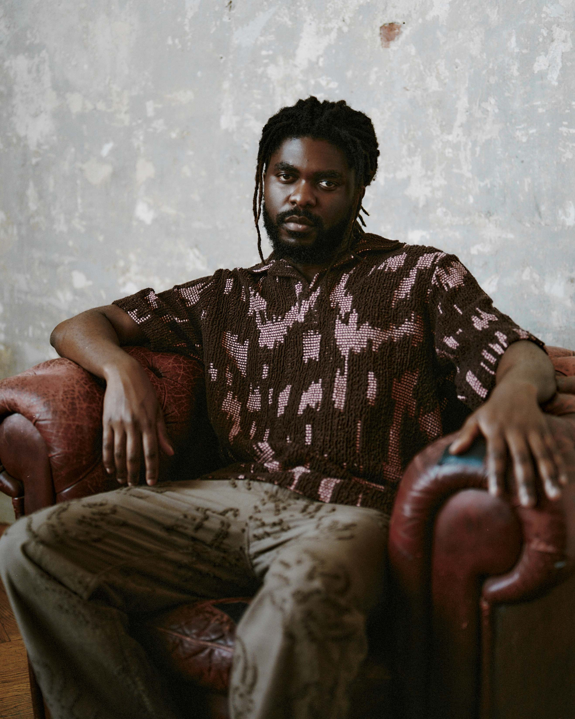 Model in a textured brown crochet shirt and embroidered trousers, seated in a leather armchair against a textured wall.