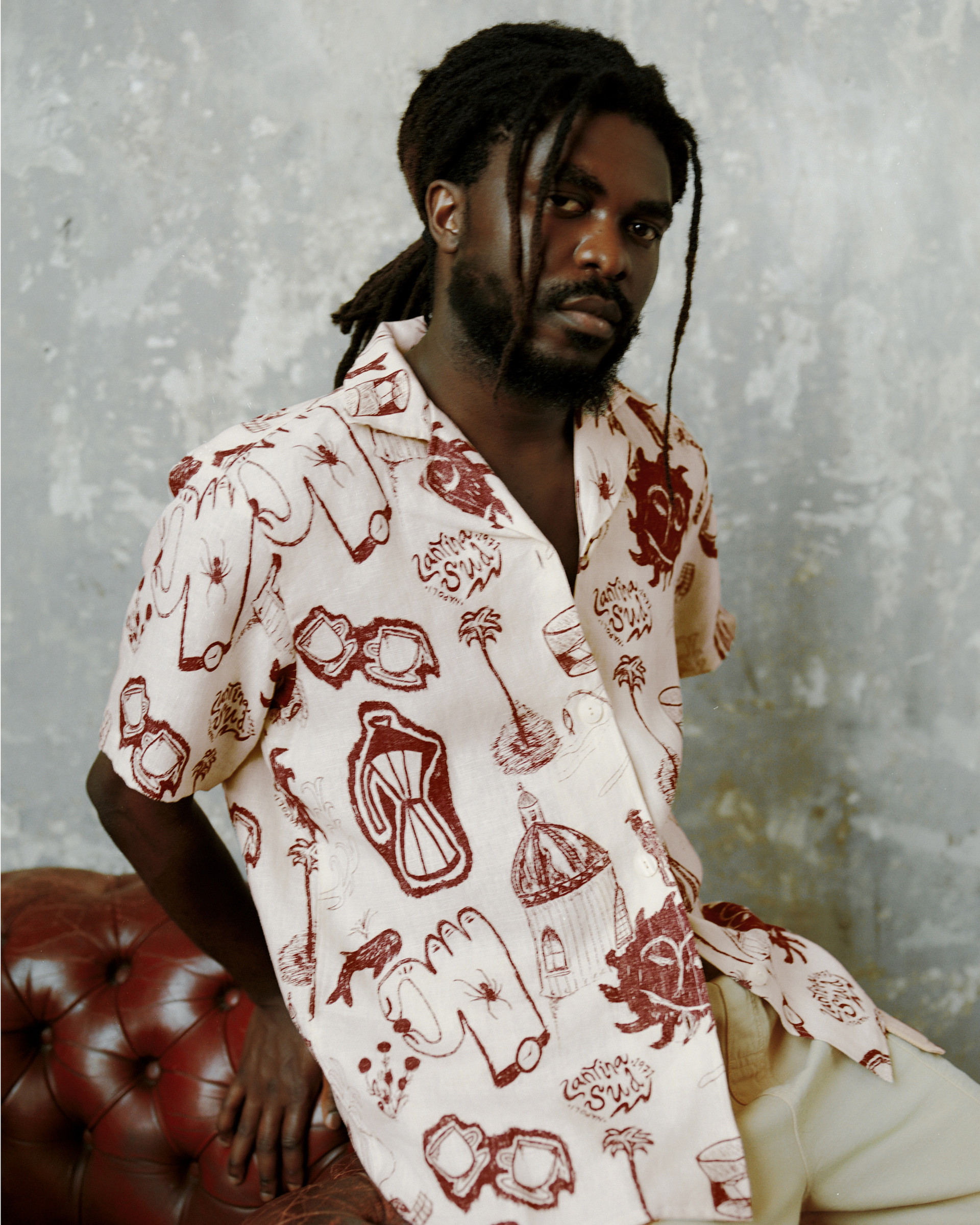 Model in a printed short-sleeve shirt with hand-drawn motifs, paired with light trousers against a textured wall.