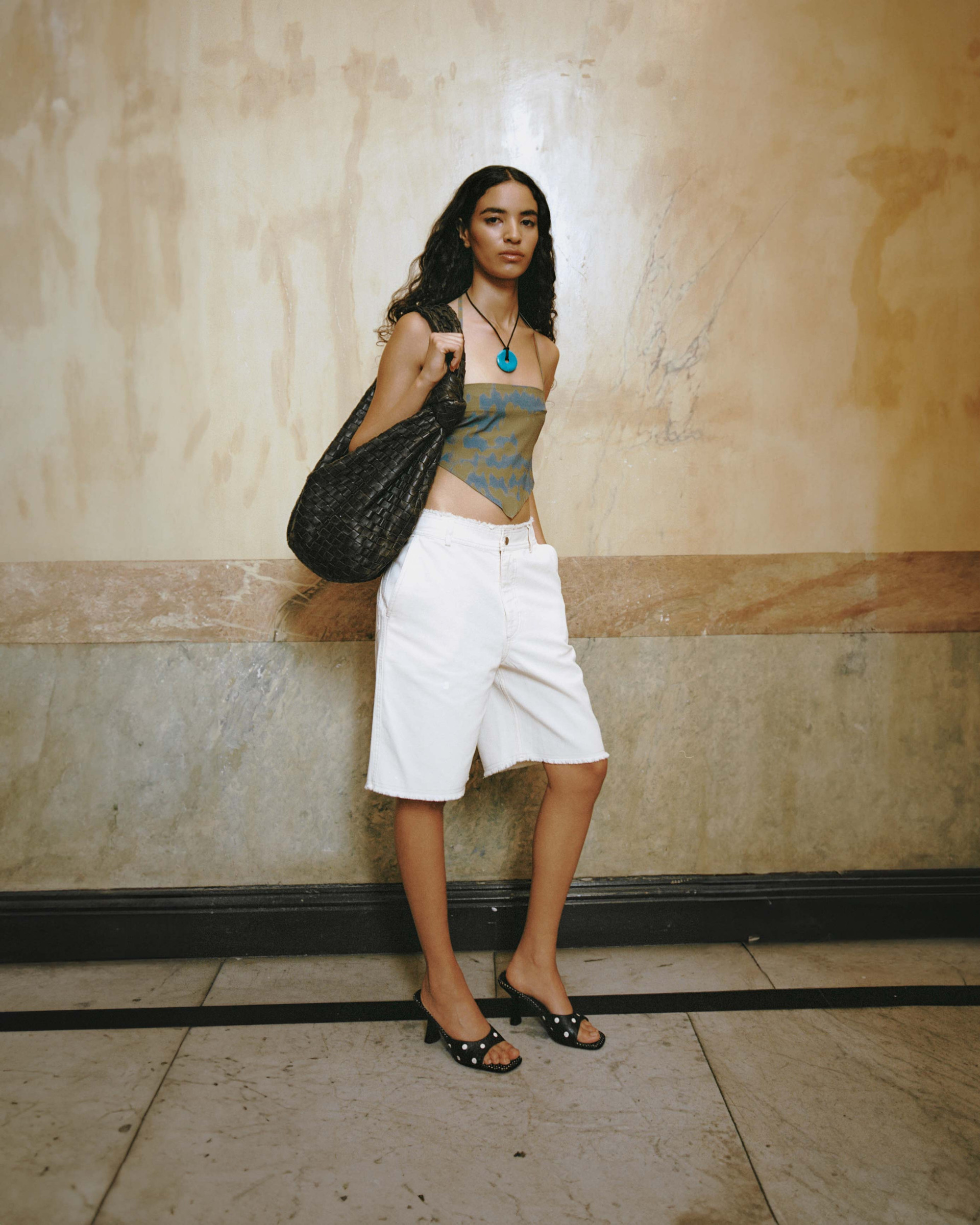 Model in a printed bandana top, frayed white knee-length shorts, black studded heels, and a woven tote against a marble wall.