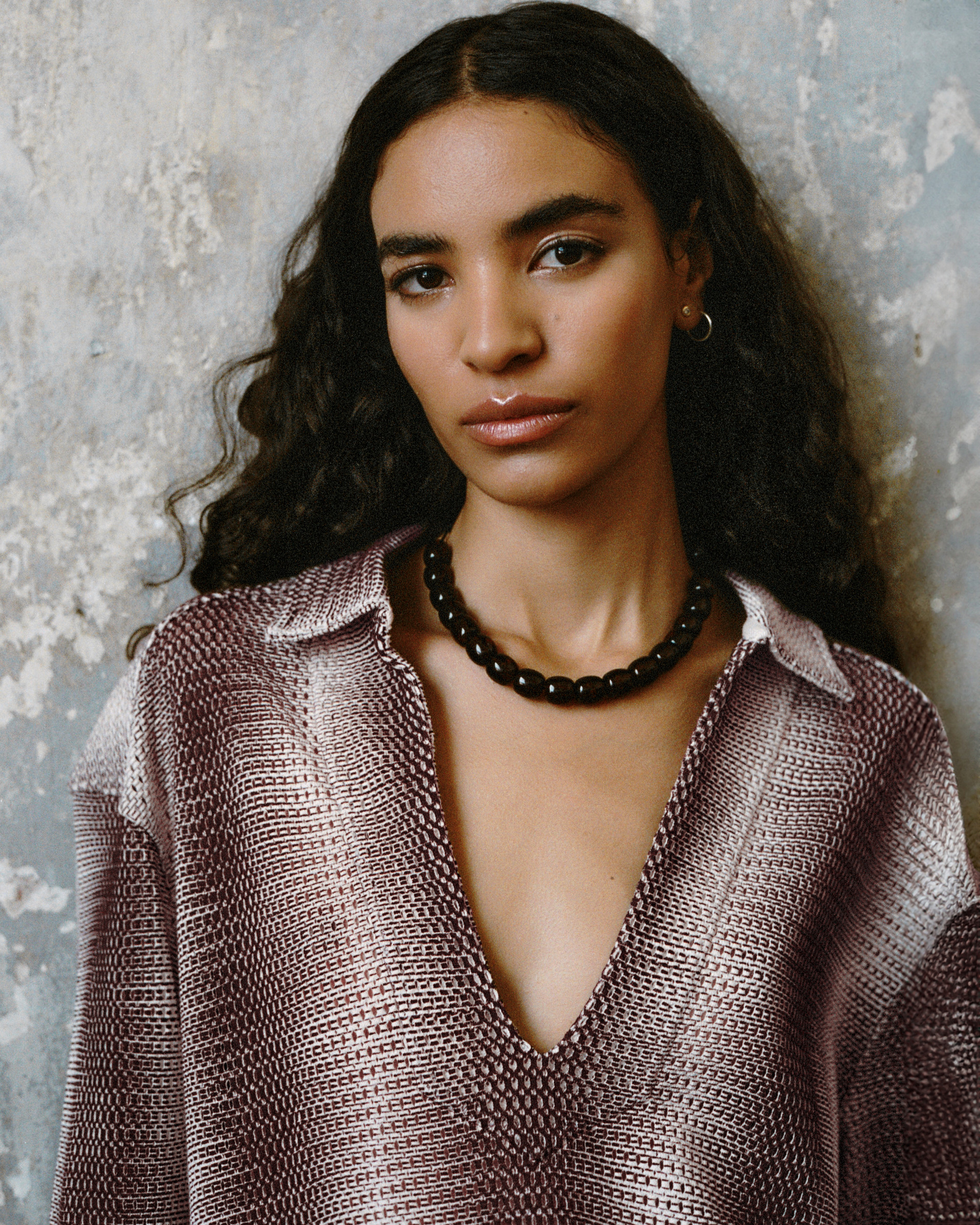 Model in a gradient knit dress with a deep neckline, paired with a black beaded necklace against a textured wall