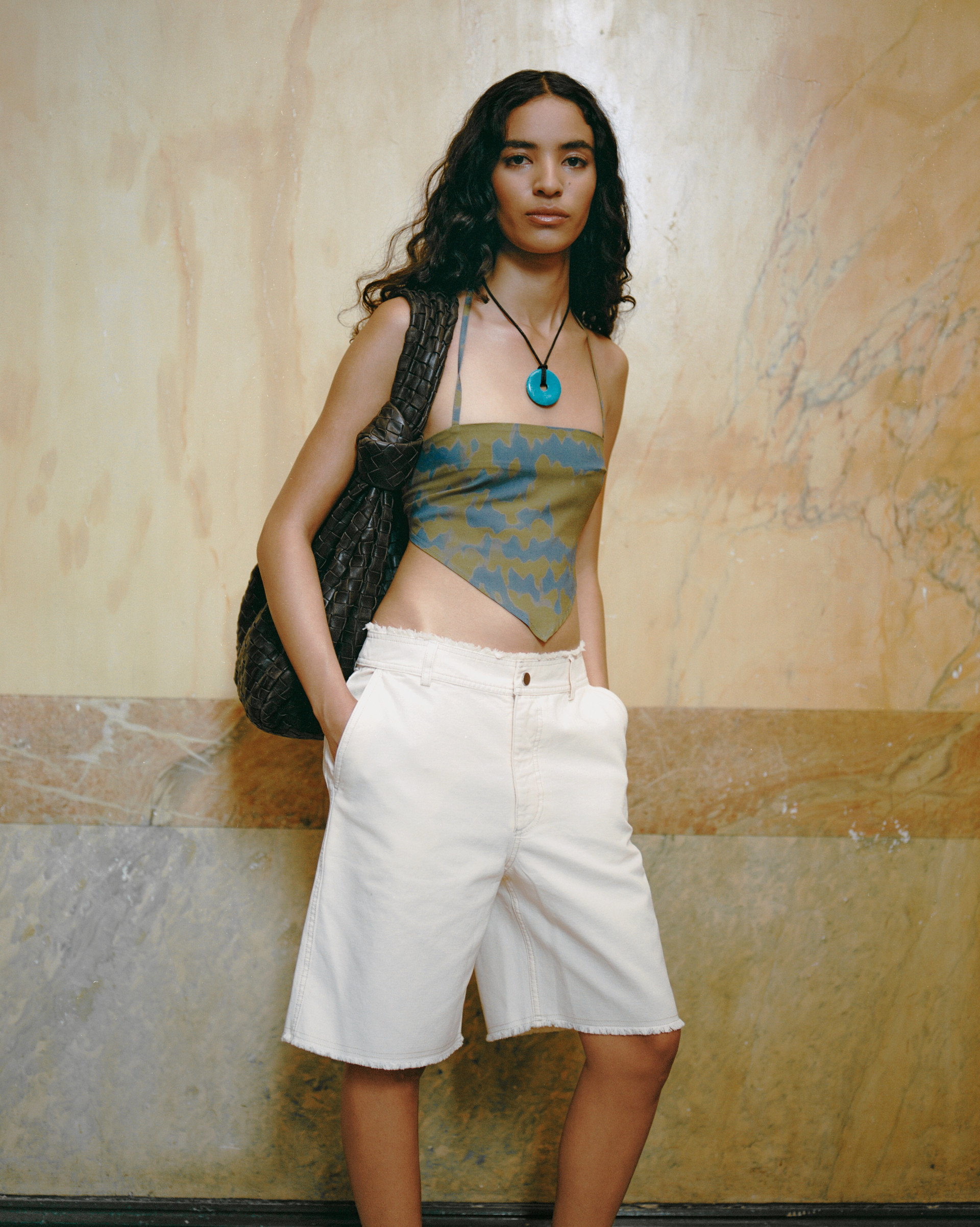 Model in a printed bandana top, frayed white knee-length shorts, a turquoise pendant, and a woven tote against a marble wall.