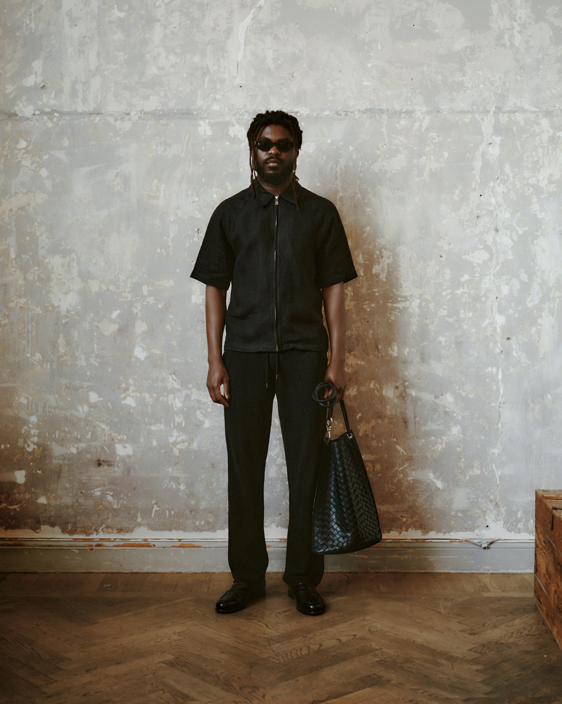 Model in an all-black outfit - zip-up short-sleeve shirt, relaxed trousers, and a woven leather tote against a textured wall.