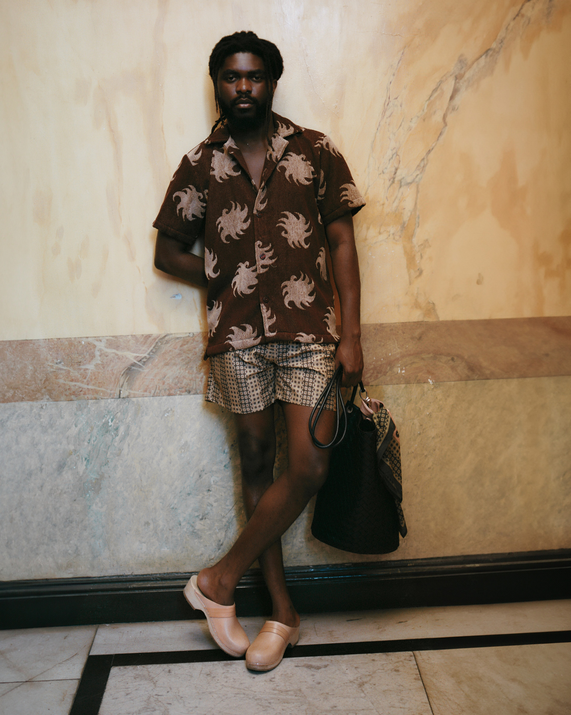 Model in a jacquard short-sleeve shirt with sun motifs, patterned swim shorts, leather clogs, and a tote against a marble wall.